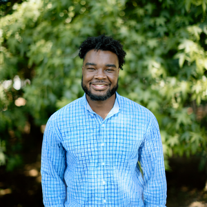 Smiling man with curly hair and beard wearing a blue checkered shirt outdoors with green trees in the background.