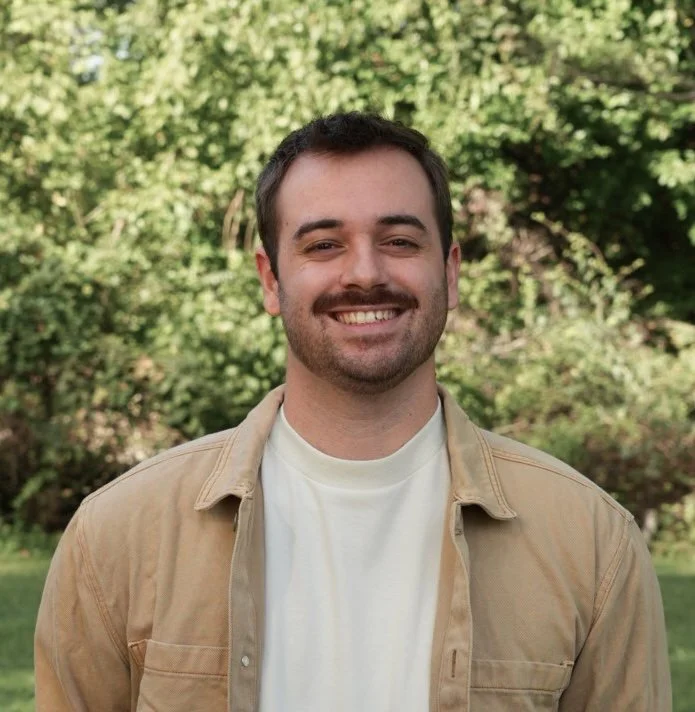 A man with a beard and short hair smiling, standing outdoors in front of green foliage, wearing a light-colored short-sleeve shirt.