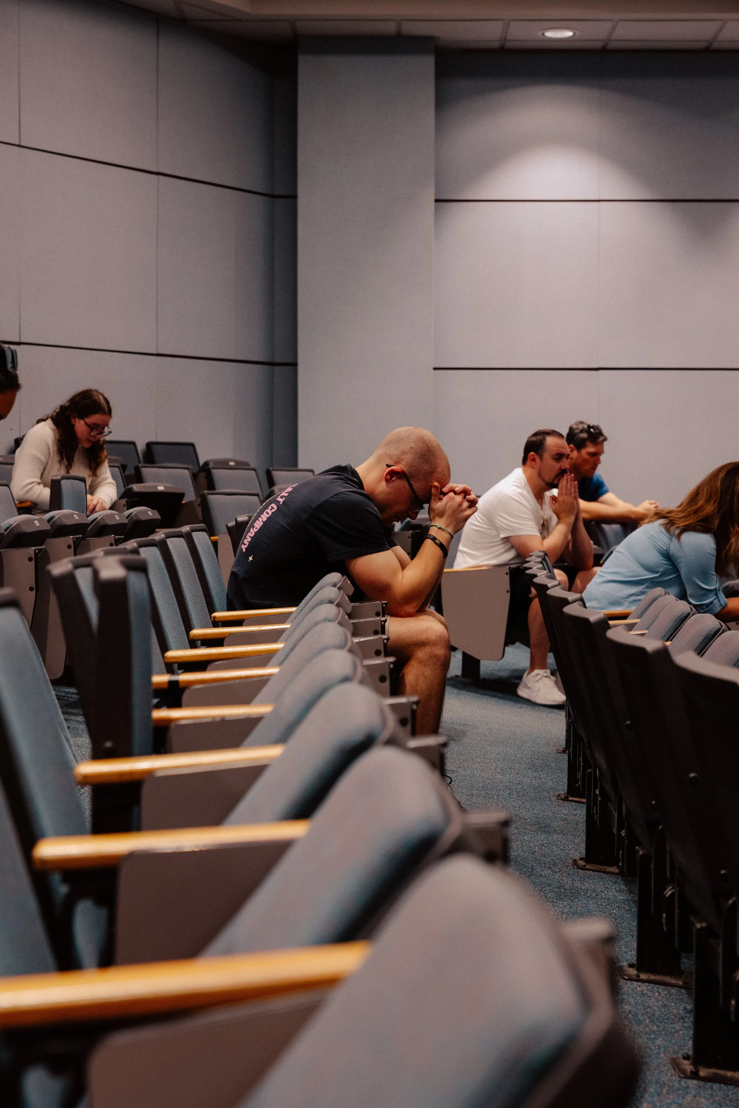 People sitting in an auditorium, some with their hands clasped in prayer or contemplation, appearing to be praying or reflecting.