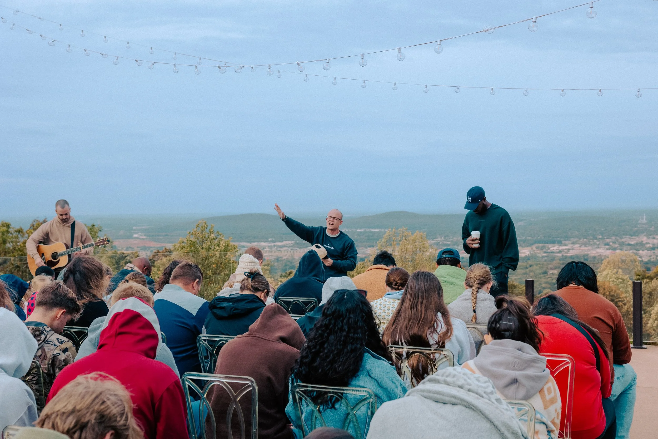 Outdoor gathering with a speaker and a musician, audience facing a scenic view with trees and hills, string lights overhead.