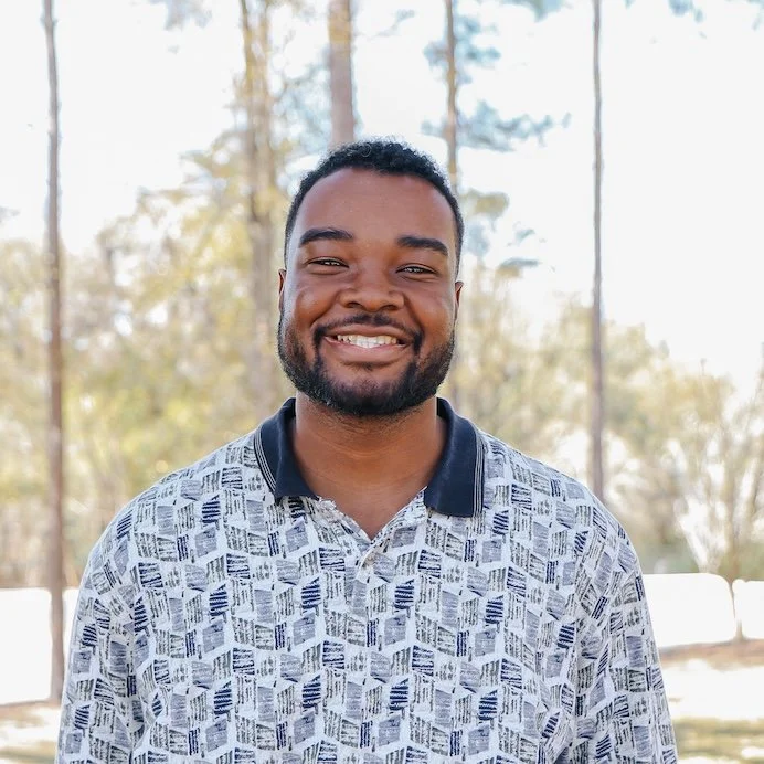 Smiling man with curly hair and beard wearing a blue checkered shirt outdoors with green trees in the background.