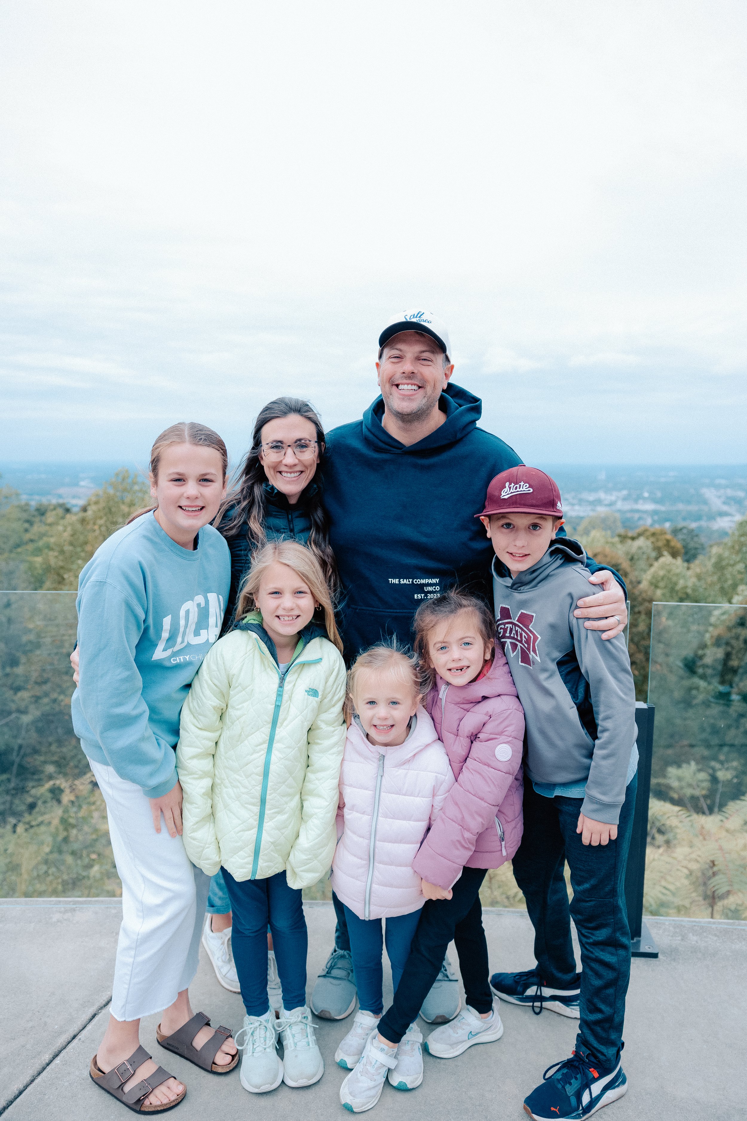 Family of seven smiling for a group photo outdoors on a cloudy day with trees and a city view in the background