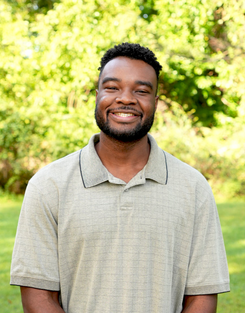 Smiling man with curly hair and beard wearing a blue checkered shirt outdoors with green trees in the background.