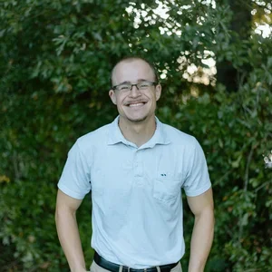 A smiling man wearing glasses and a light blue polo shirt standing outdoors in front of green foliage.