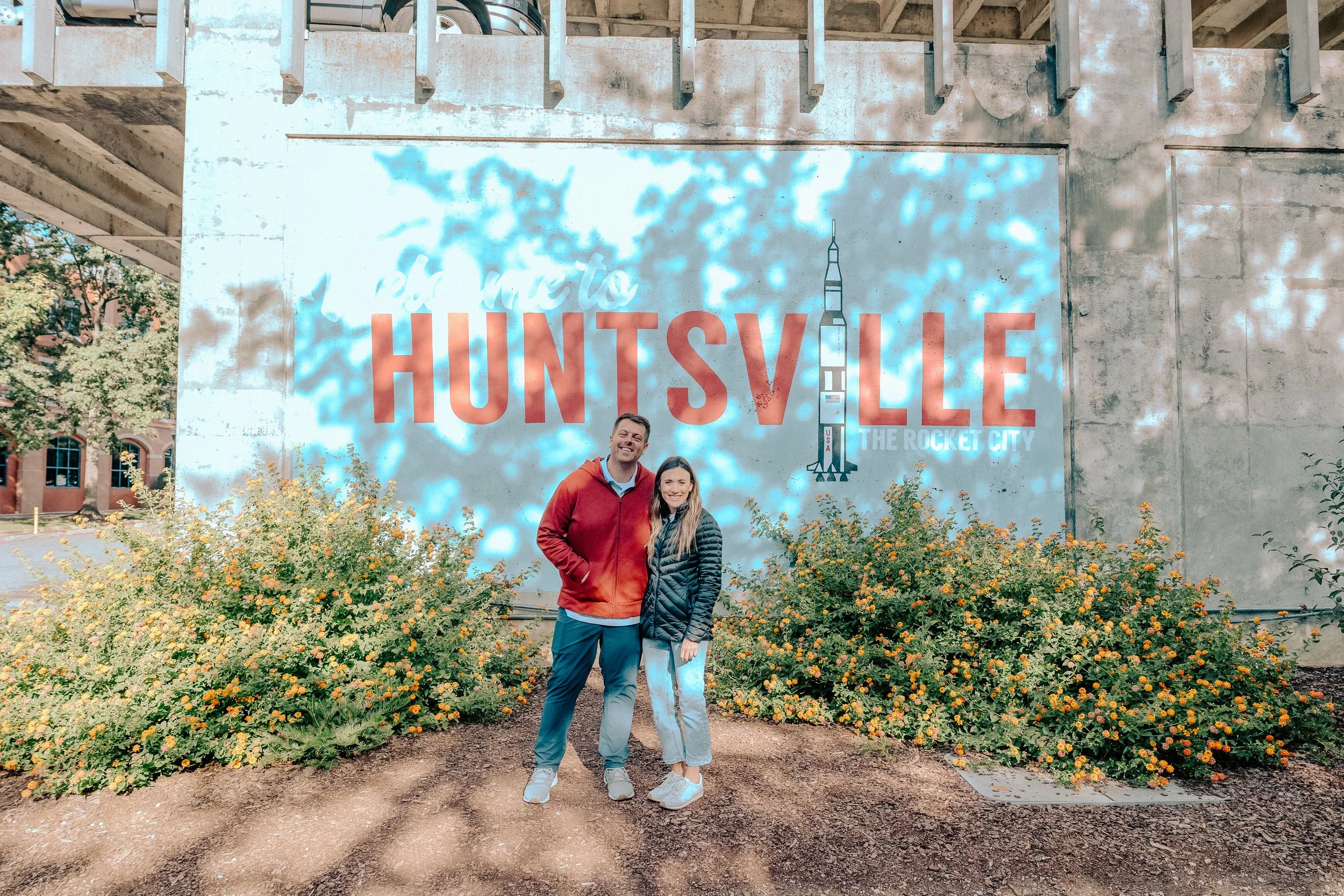 A man and a woman standing together in front of a large wall with an illustration of a rocket and the text "Welcome to Huntsville, the Rocket City." They are smiling, with bushes with yellow flowers at their feet, under a structure casting dappled sh
