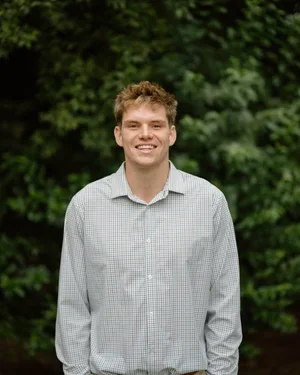 Young man smiling outdoors with green trees in the background.