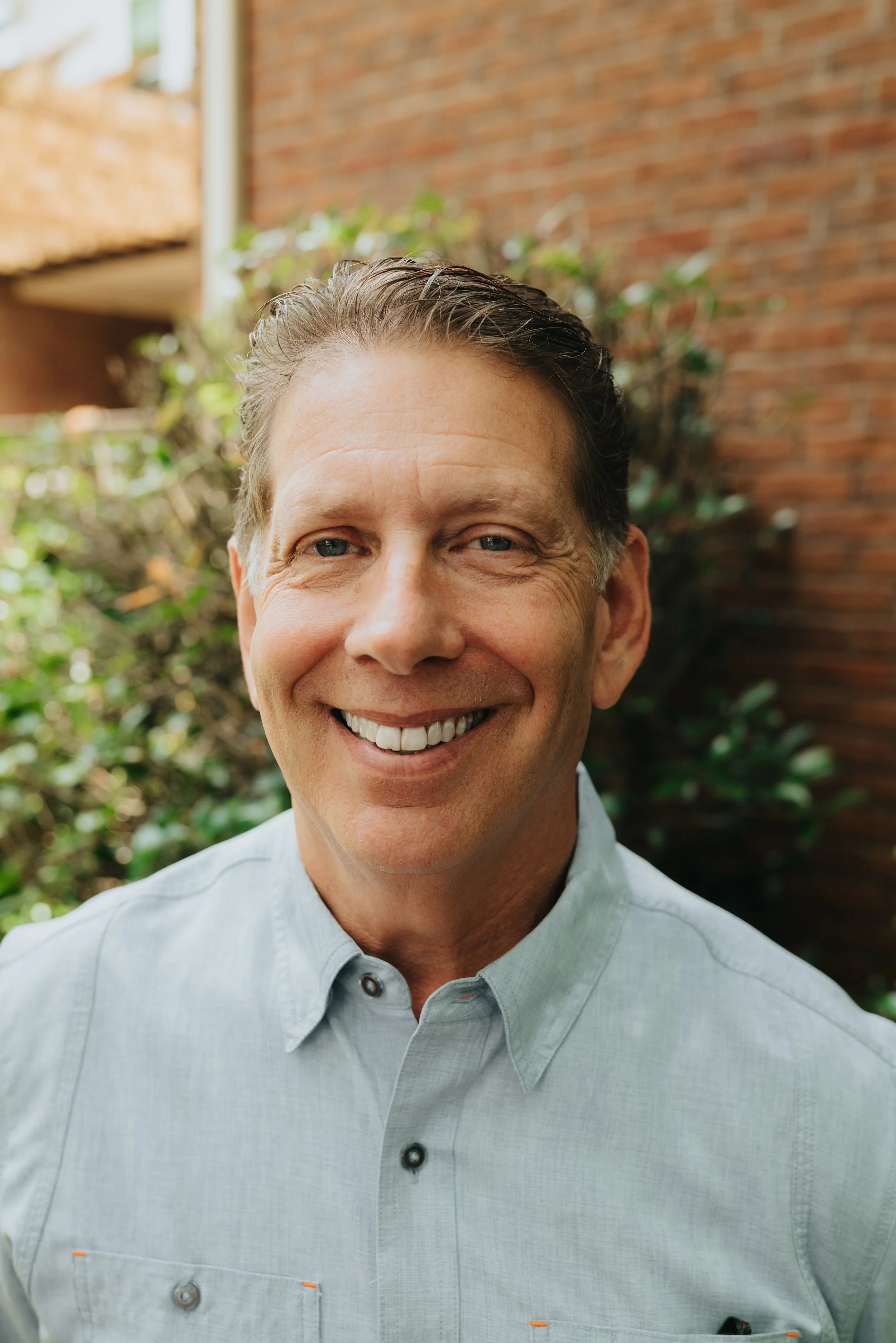 A smiling man standing outdoors in front of trees, wearing a blue button-up shirt.