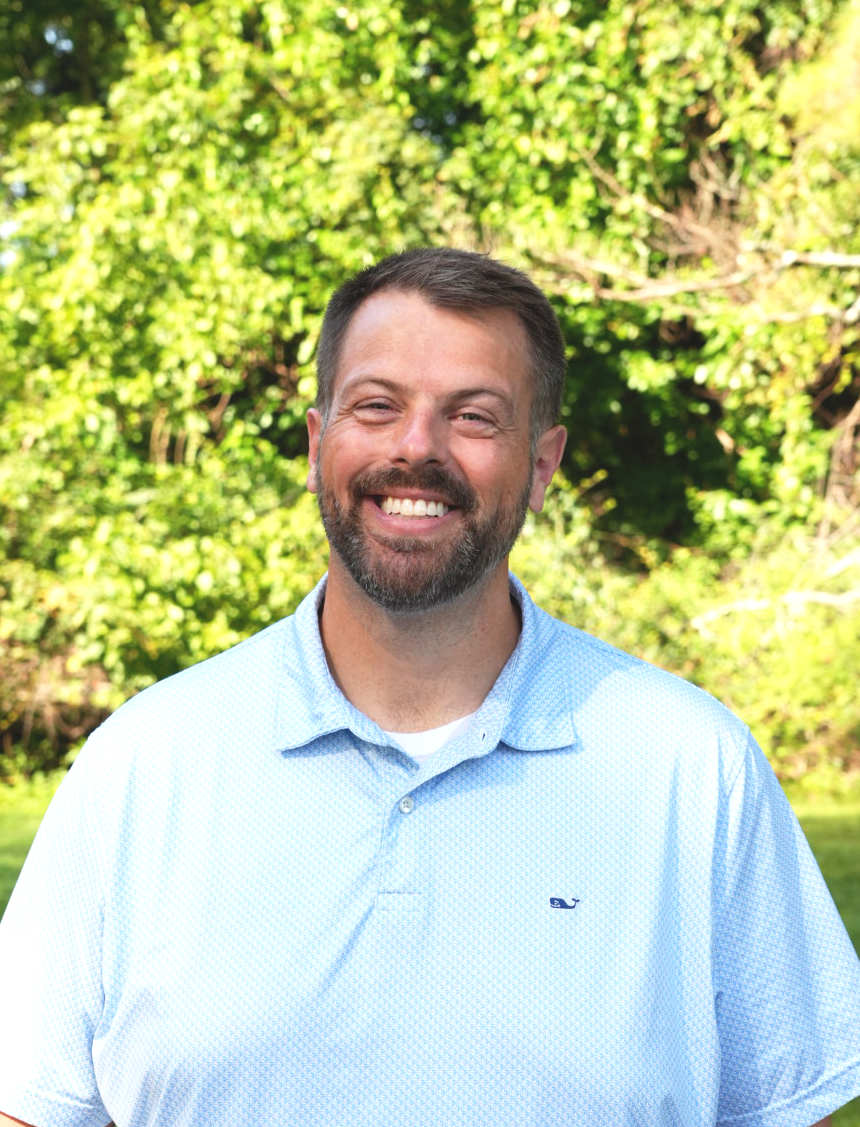 A smiling man standing outdoors in front of trees, wearing a blue button-up shirt.
