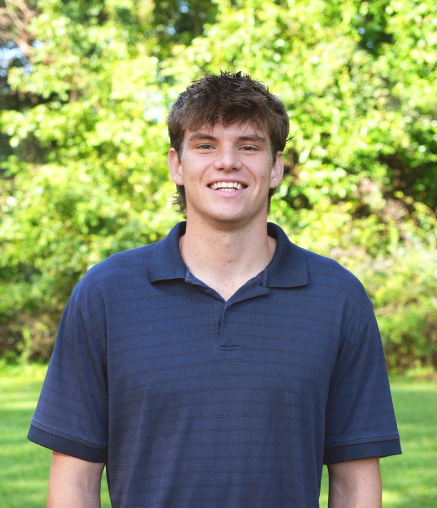 Young man smiling outdoors with green trees in the background.