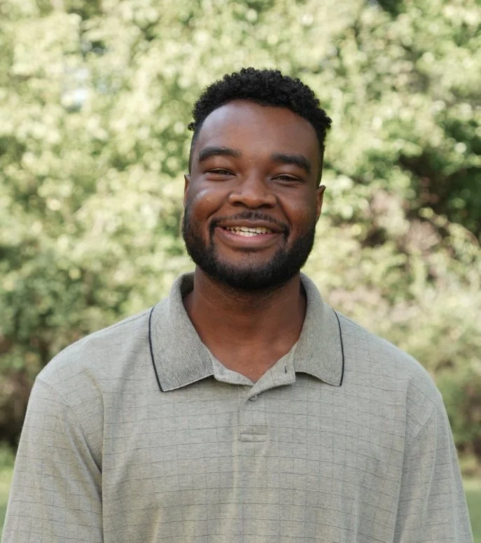 Smiling man with curly hair and beard wearing a blue checkered shirt outdoors with green trees in the background.