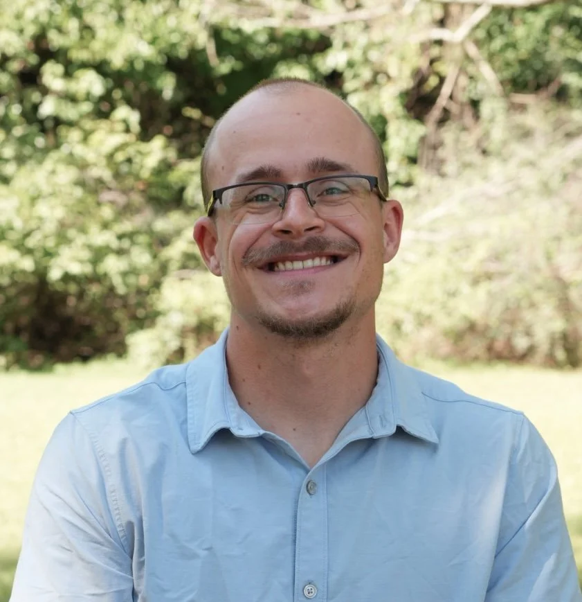 A smiling man wearing glasses and a light blue polo shirt standing outdoors in front of green foliage.