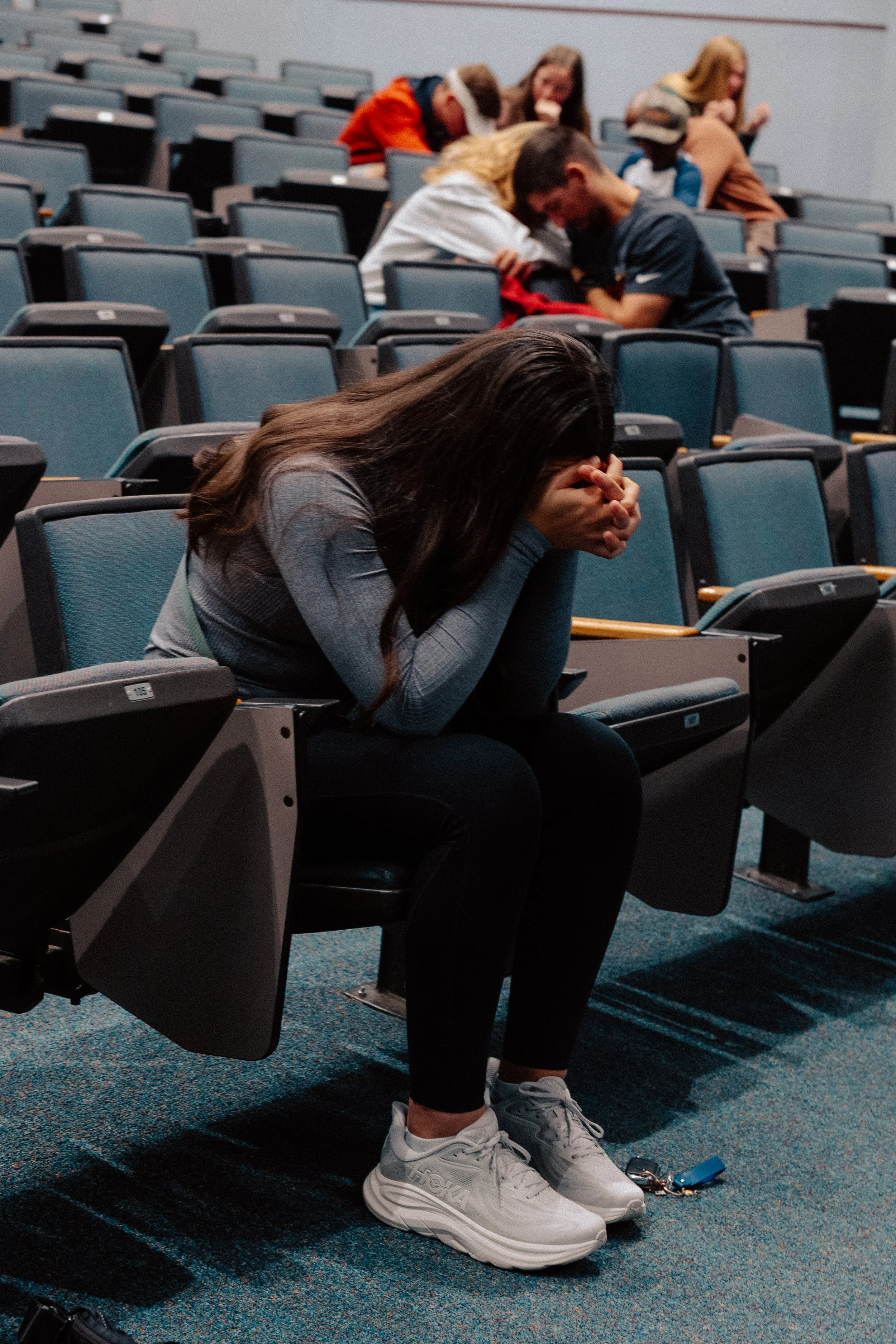 A young woman sitting alone in an auditorium, covering her face with her hands, surrounded by empty seats and other people in the background.