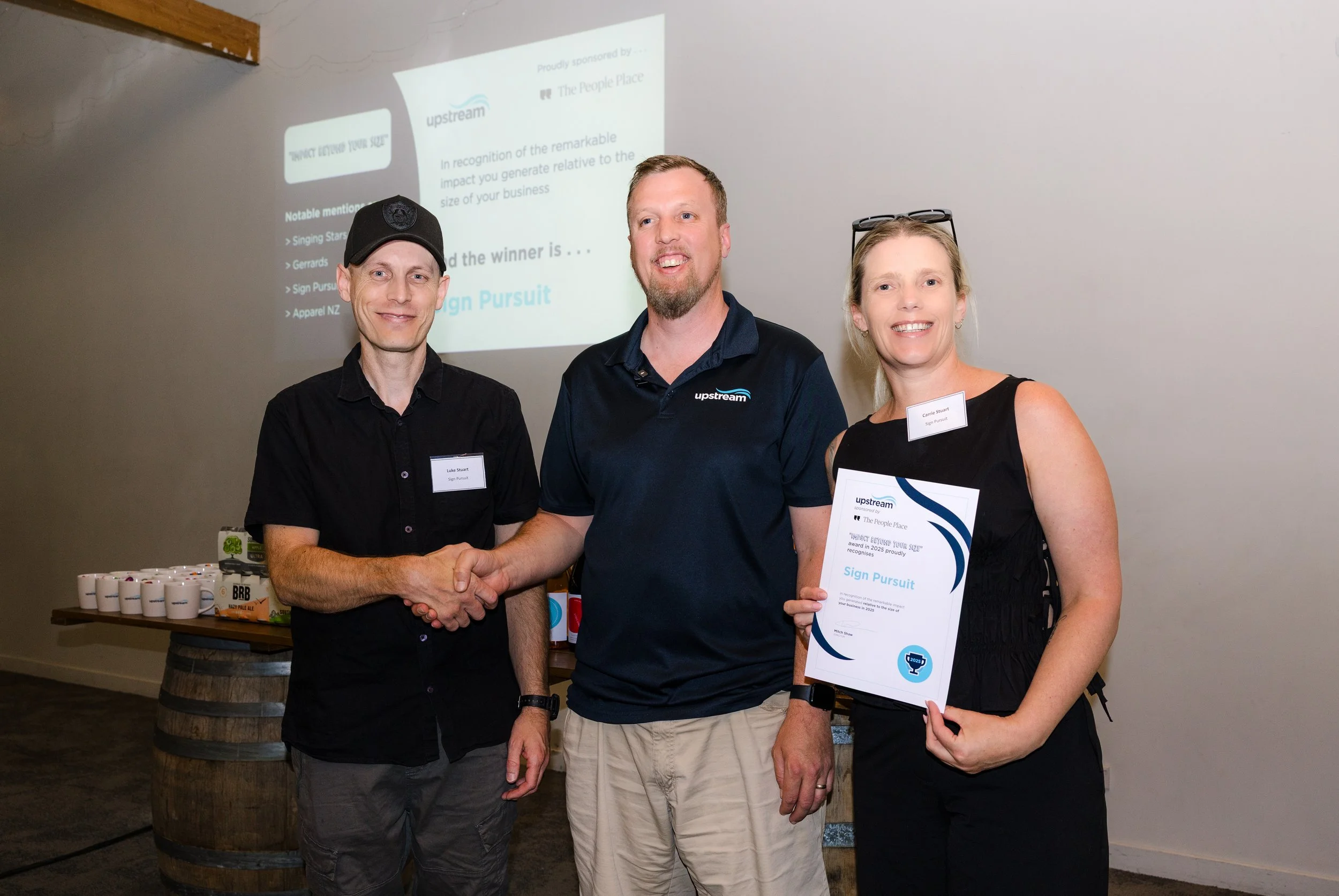Three people standing indoors, two men and one woman, shaking hands, with a man in a black shirt and woman holding a certificate. In the background, a projected presentation slide, and a table with coffee mugs and snacks.