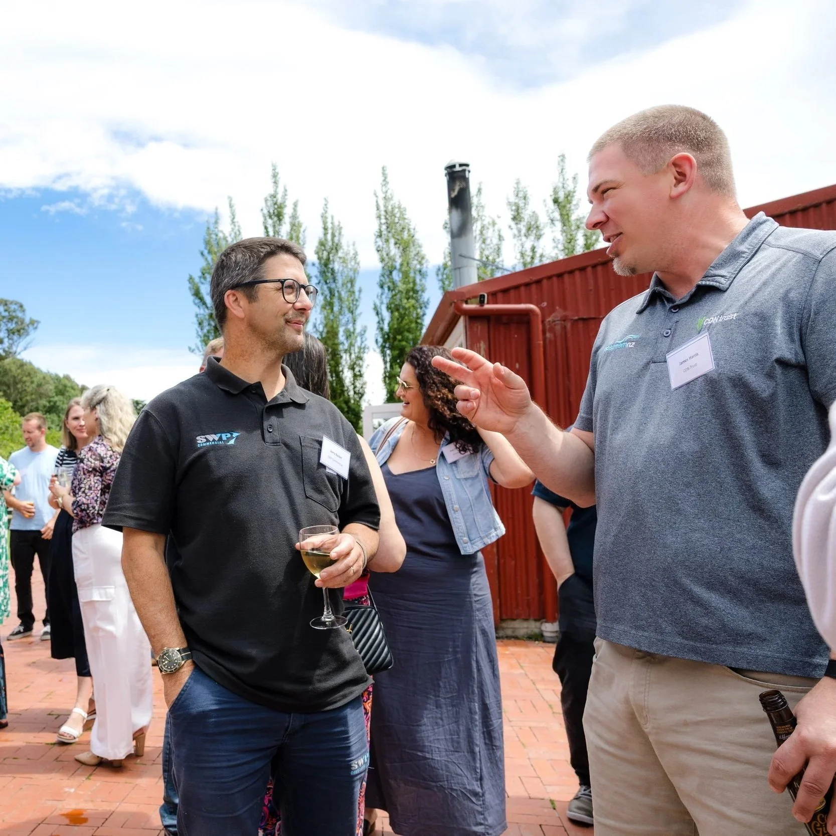 Two men are in conversation at an outdoor gathering, with a group of people in the background, some holding drinks, under a partly cloudy sky.