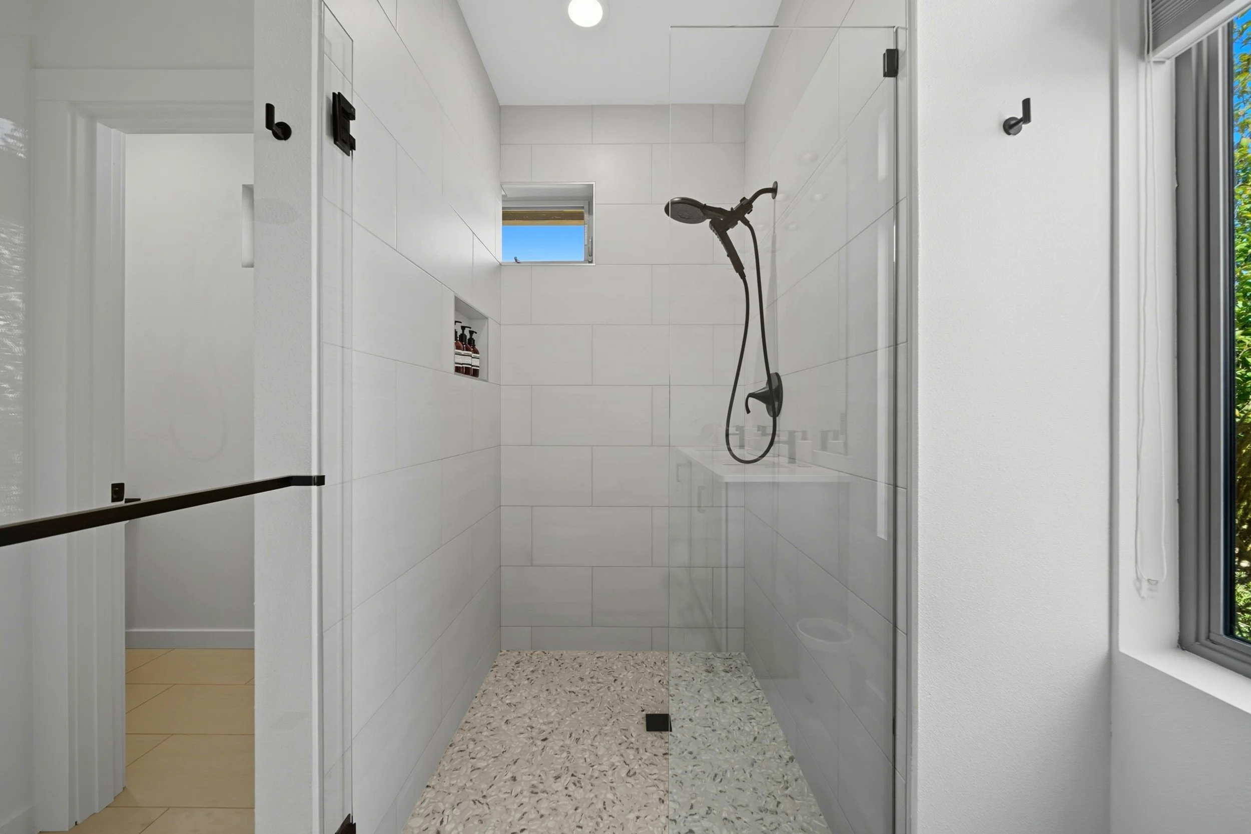 Modern shower with white tiled walls, a small window, and personal care products on a niche. The shower has a black handheld showerhead and a glass door.