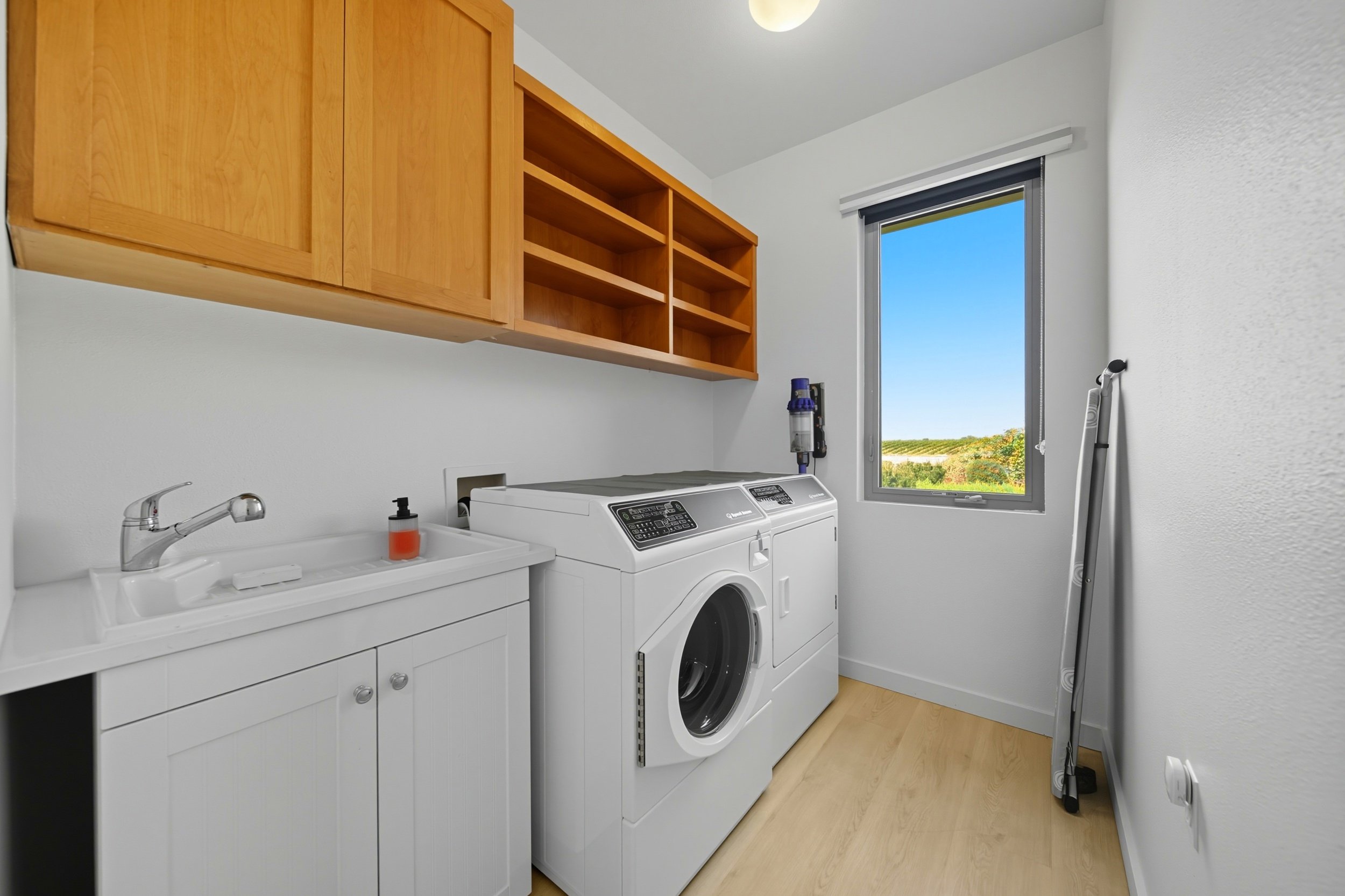 A laundry room with a white washer, a sink with soap, wooden cabinets, a window with a view of greenery, and an iron leaning against the wall.