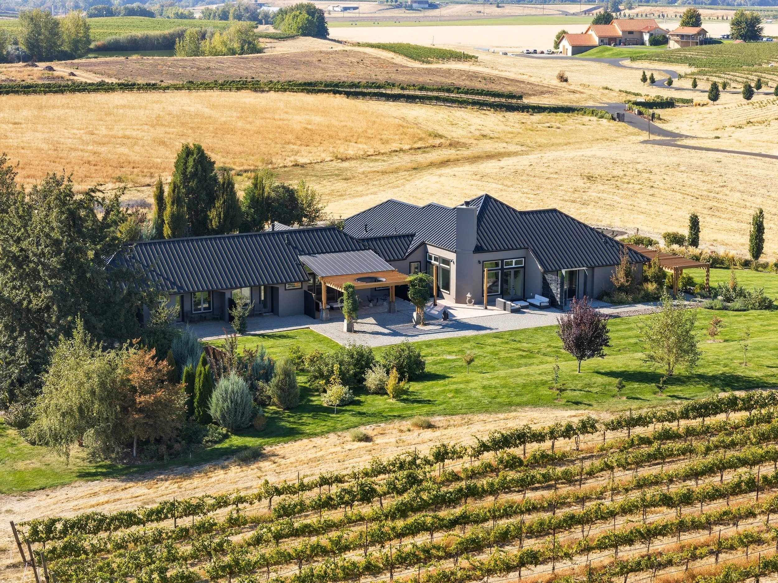 A modern ranch-style house with a black metal roof, large windows, and outdoor patios, surrounded by landscaped yard and trees, overlooking rolling vineyards and farmland.