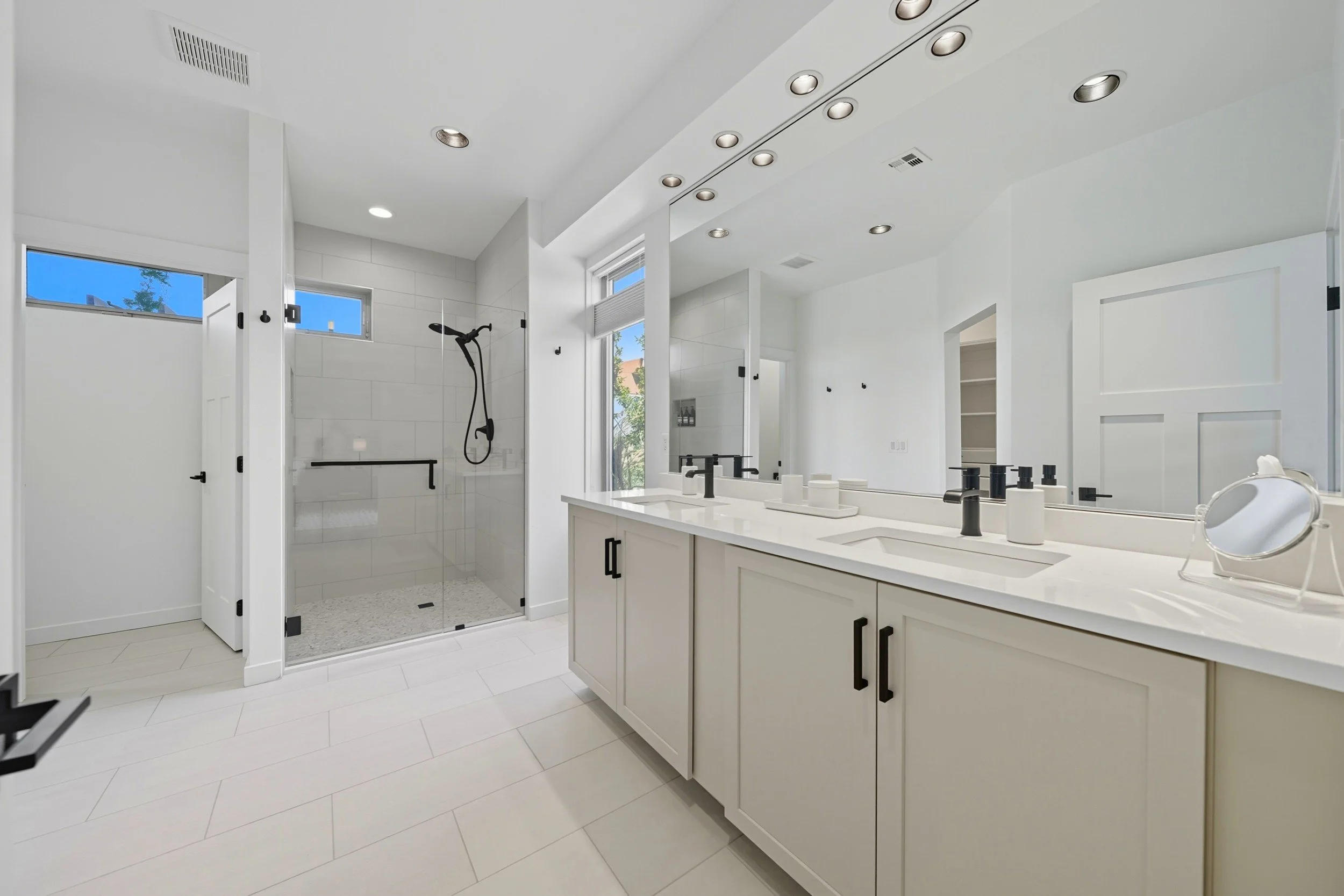 Modern white bathroom with dual sinks, large mirror, glass-enclosed shower with black fixtures, small windows, and minimalist decor.