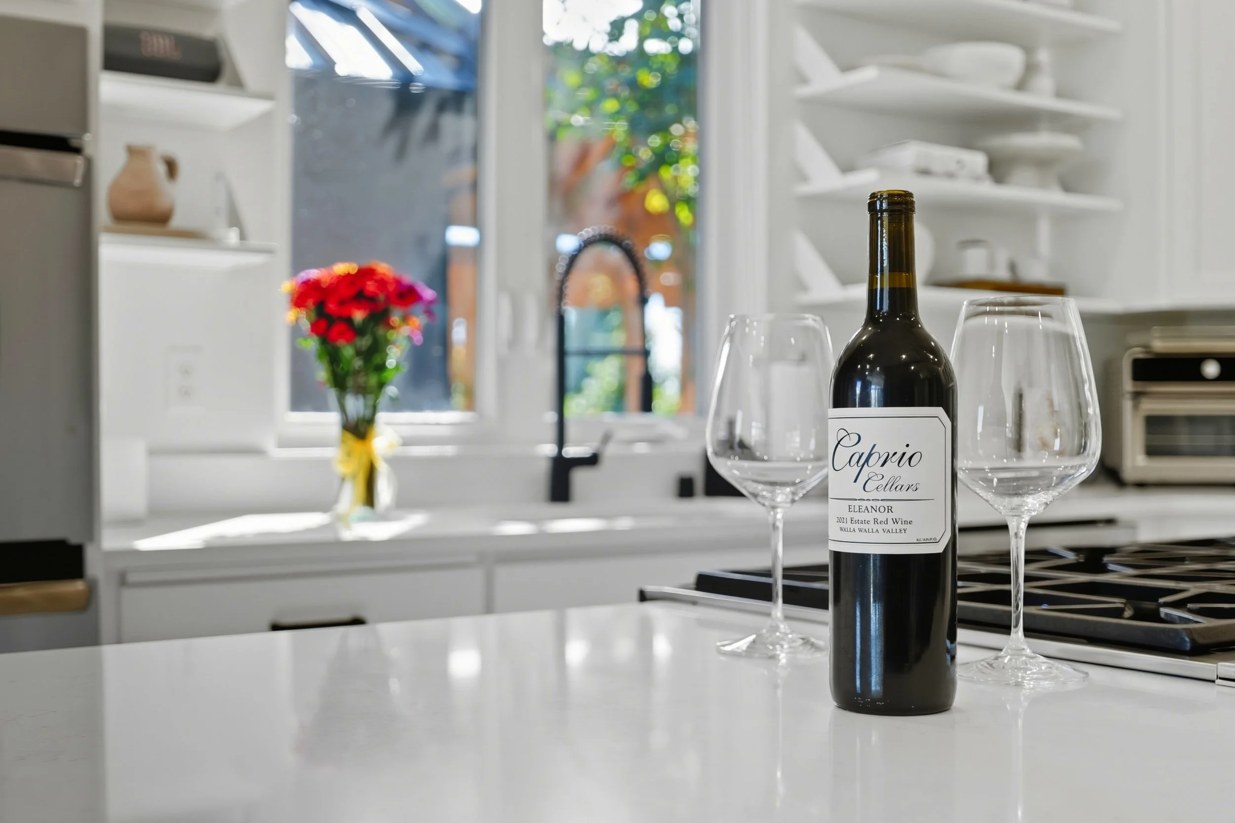 A bottle of red wine and two empty wine glasses on a kitchen countertop, with a blurry window and a vase of red and yellow flowers in the background.