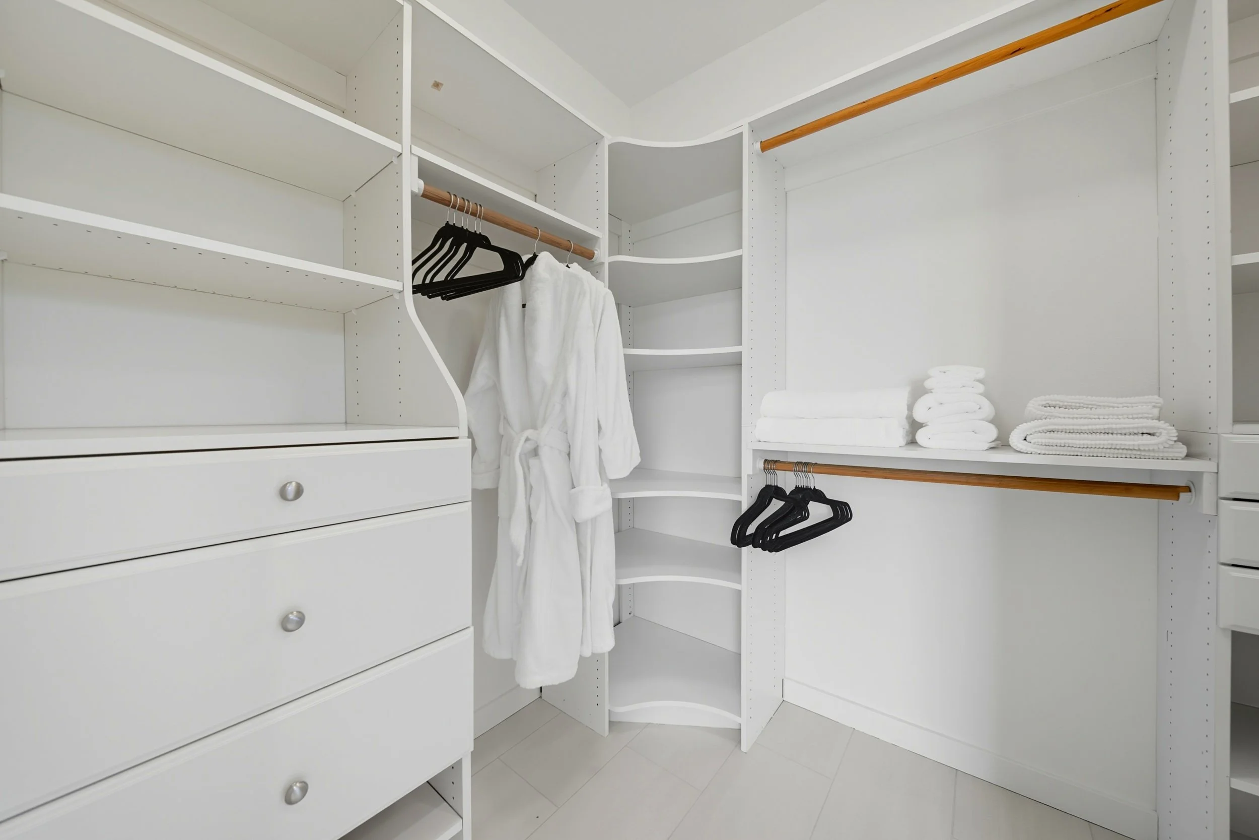 Empty white closet with shelves, hanging rods, folded towels, and a white bathrobe hanging on a hanger.