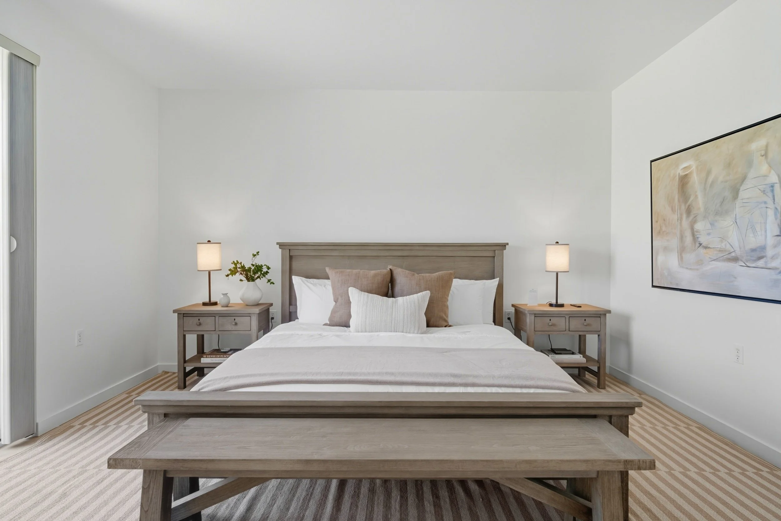 A minimalist bedroom with a large bed, beige pillows, white bedding, and wooden furniture. Two nightstands with lamps and decor, a large abstract painting on the wall, and light-colored striped carpet.
