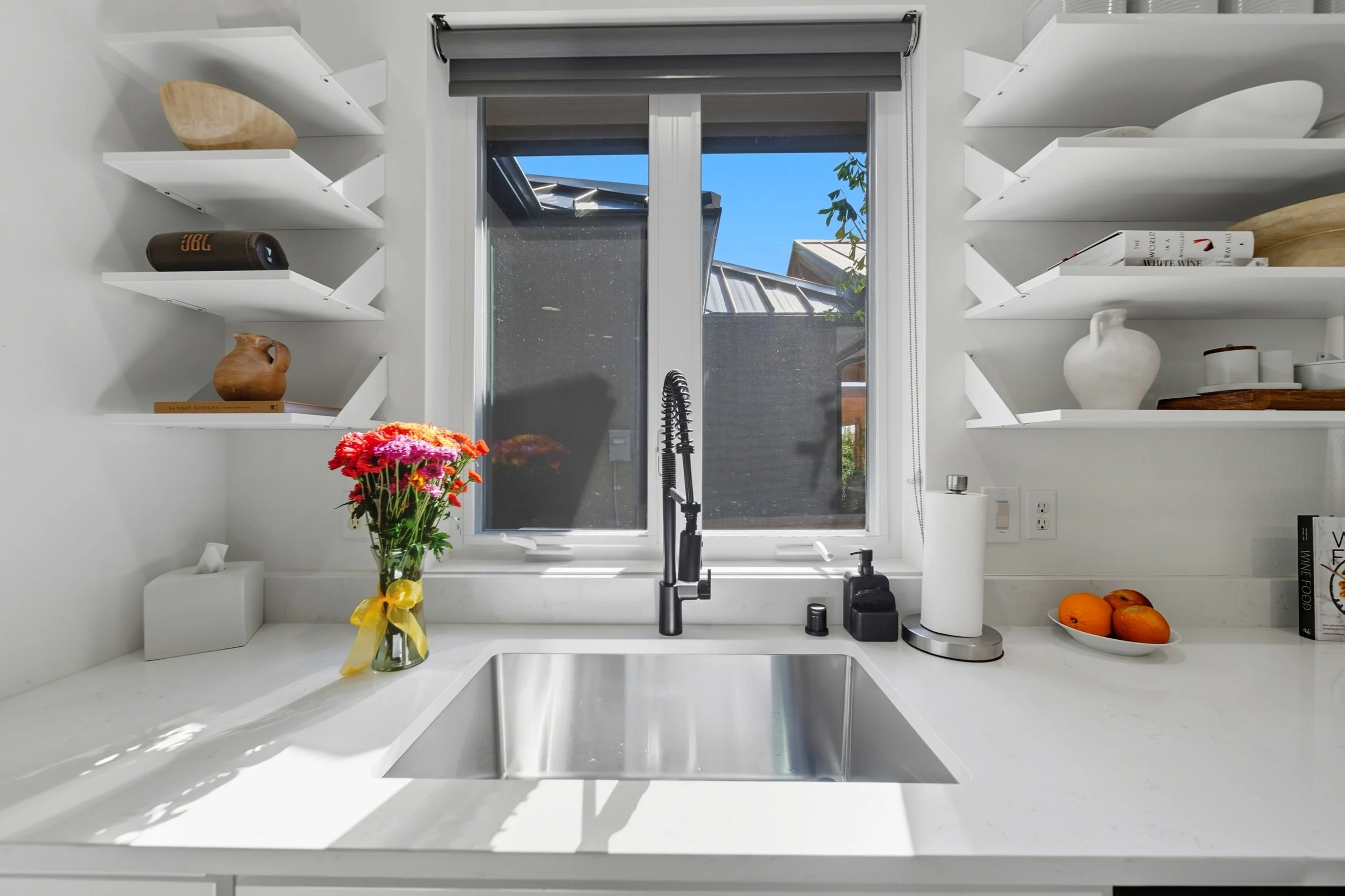 White kitchen countertop with a sink, a vase of pink and orange flowers, a roll of paper towels, and a bowl of oranges. Open white shelves with decorative items and a window with a view of a backyard.