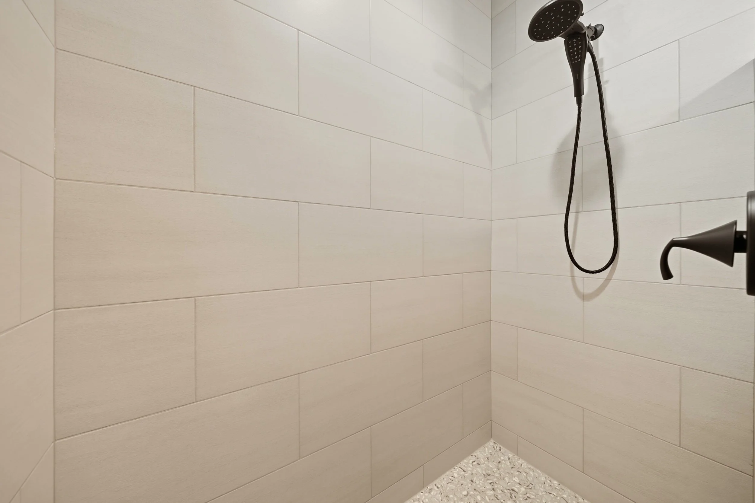 A walk-in shower with beige wall tiles, a black showerhead, and a black handle, with a textured pebble floor.