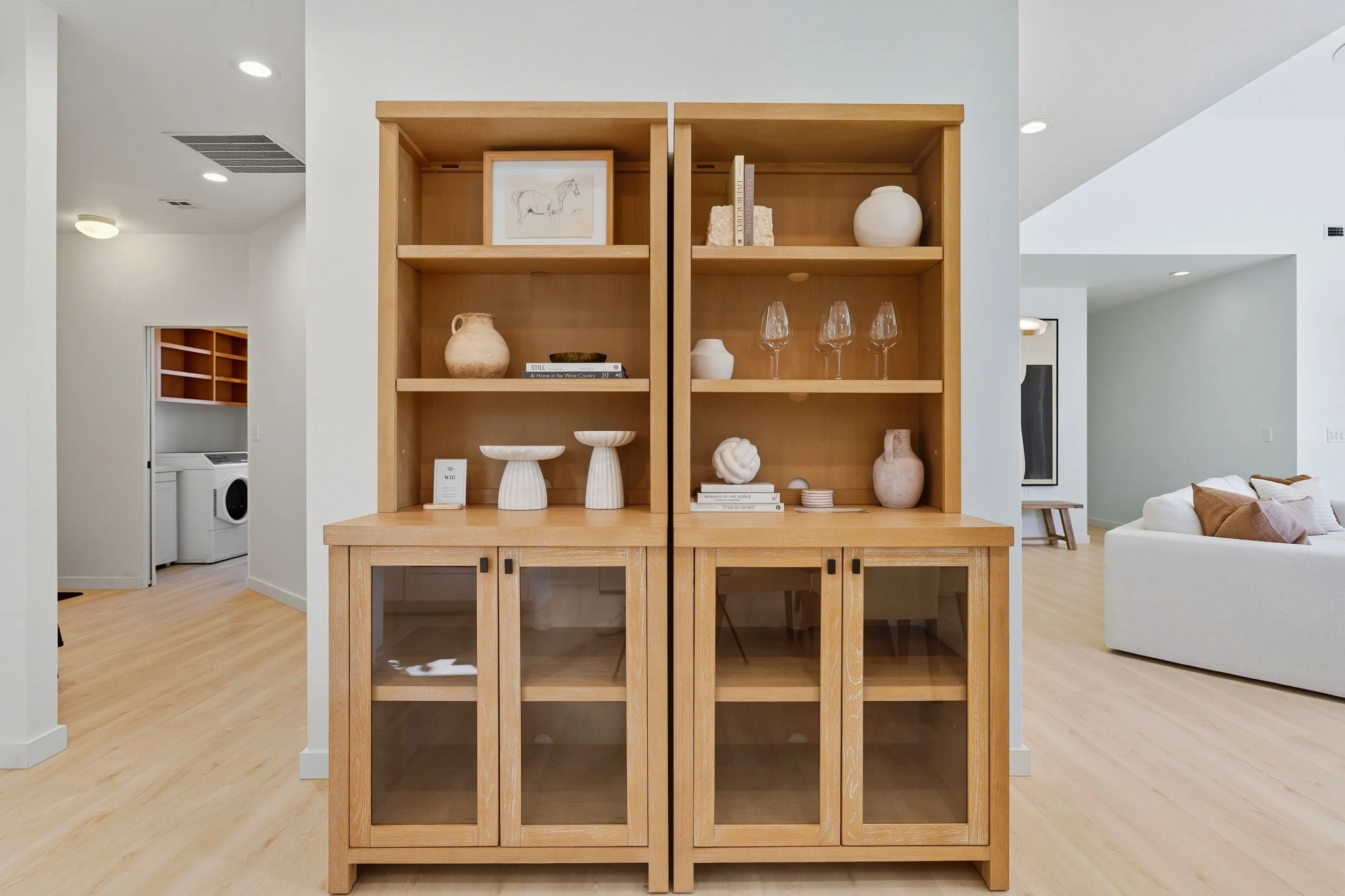 A wooden display cabinet with glass doors on the lower section, filled with decorative ceramics and books, situated in a modern living room with a light-colored wood floor and white walls.