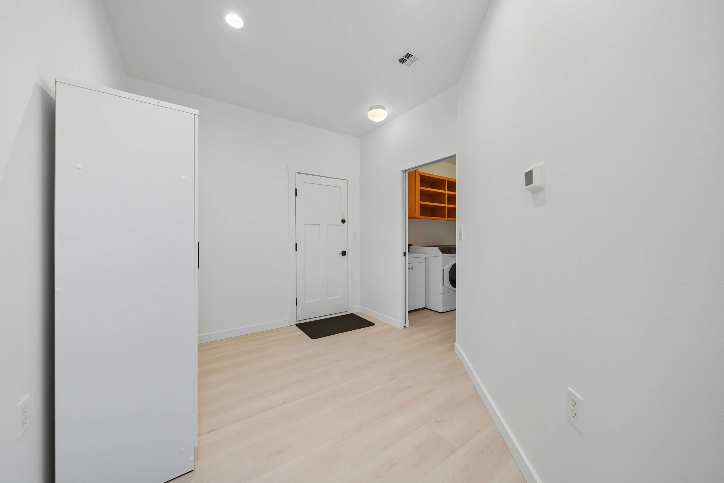 A minimalistic laundry room with white walls, a small cabinet, and an open door leading to a laundry area with a washing machine and wooden shelves.
