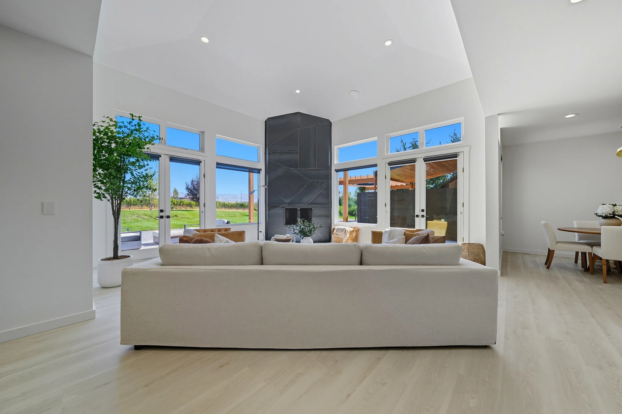 Bright living room with large windows, white walls, a white sofa, beige chairs, a stone fireplace, and a view of a green yard outside.