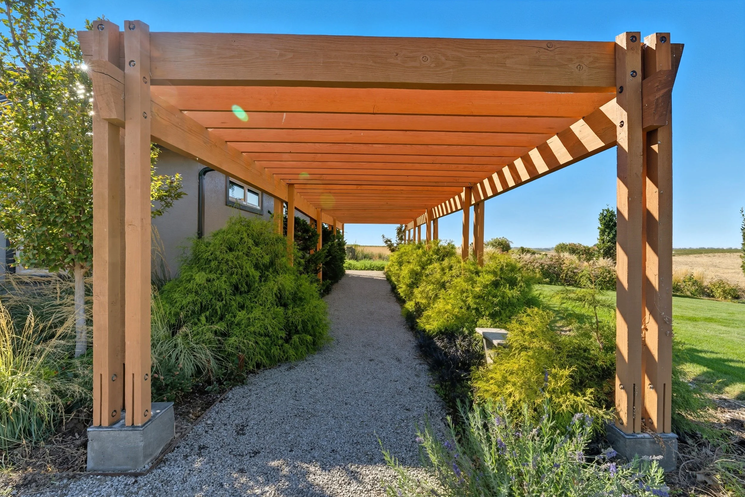 Wooden pergola over gravel pathway, surrounded by green shrubs and plants with a blue sky in the background.