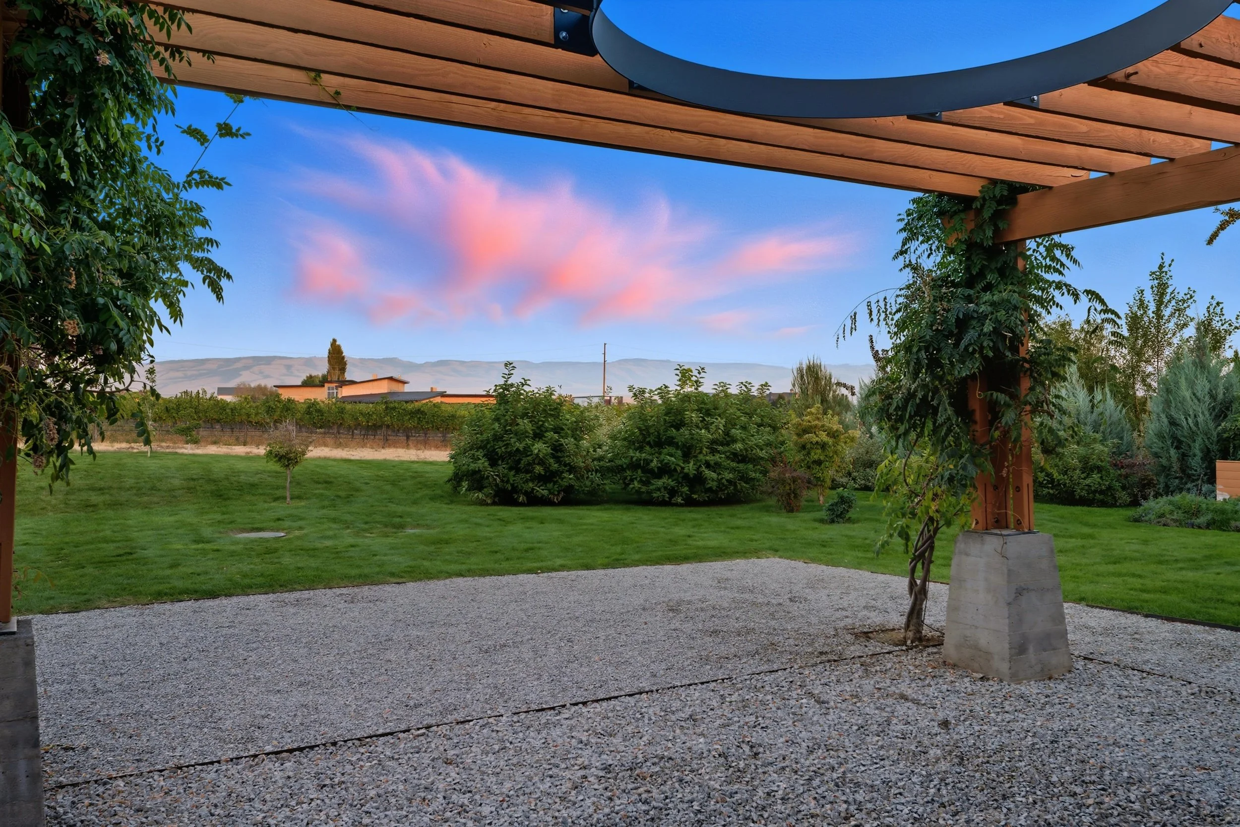 A backyard view from a patio with a gravel surface, under a wooden pergola with a circular cutout in the roof. The yard has green grass, various trees and shrubs, and a scenic background of mountains and a pink-and-blue sky during sunset.