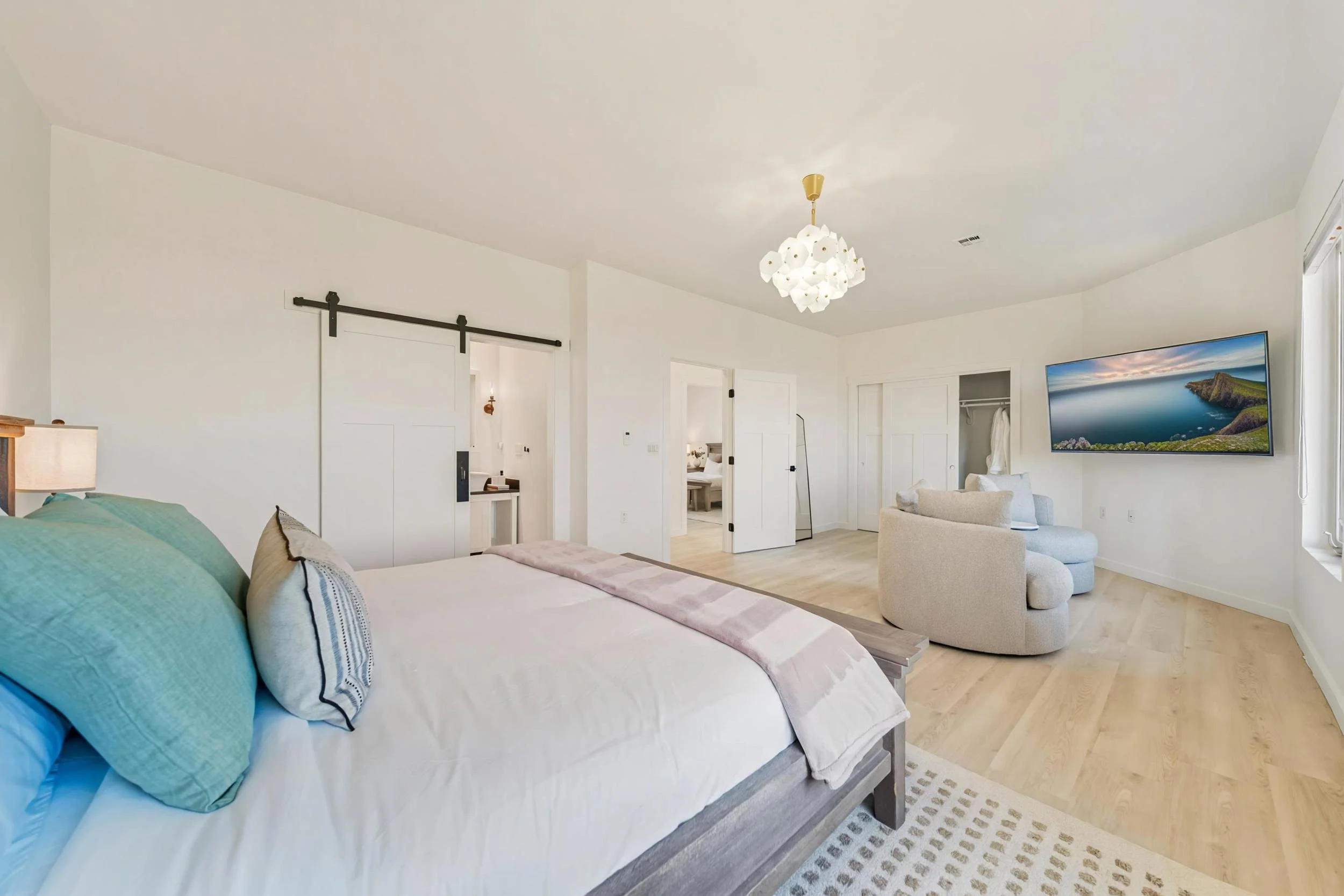 Spacious bedroom with a large bed, light-colored bedding, and decorative pillows, a sitting area with a cream armchair and a wall-mounted TV, light wood flooring, white walls, and a modern chandelier.
