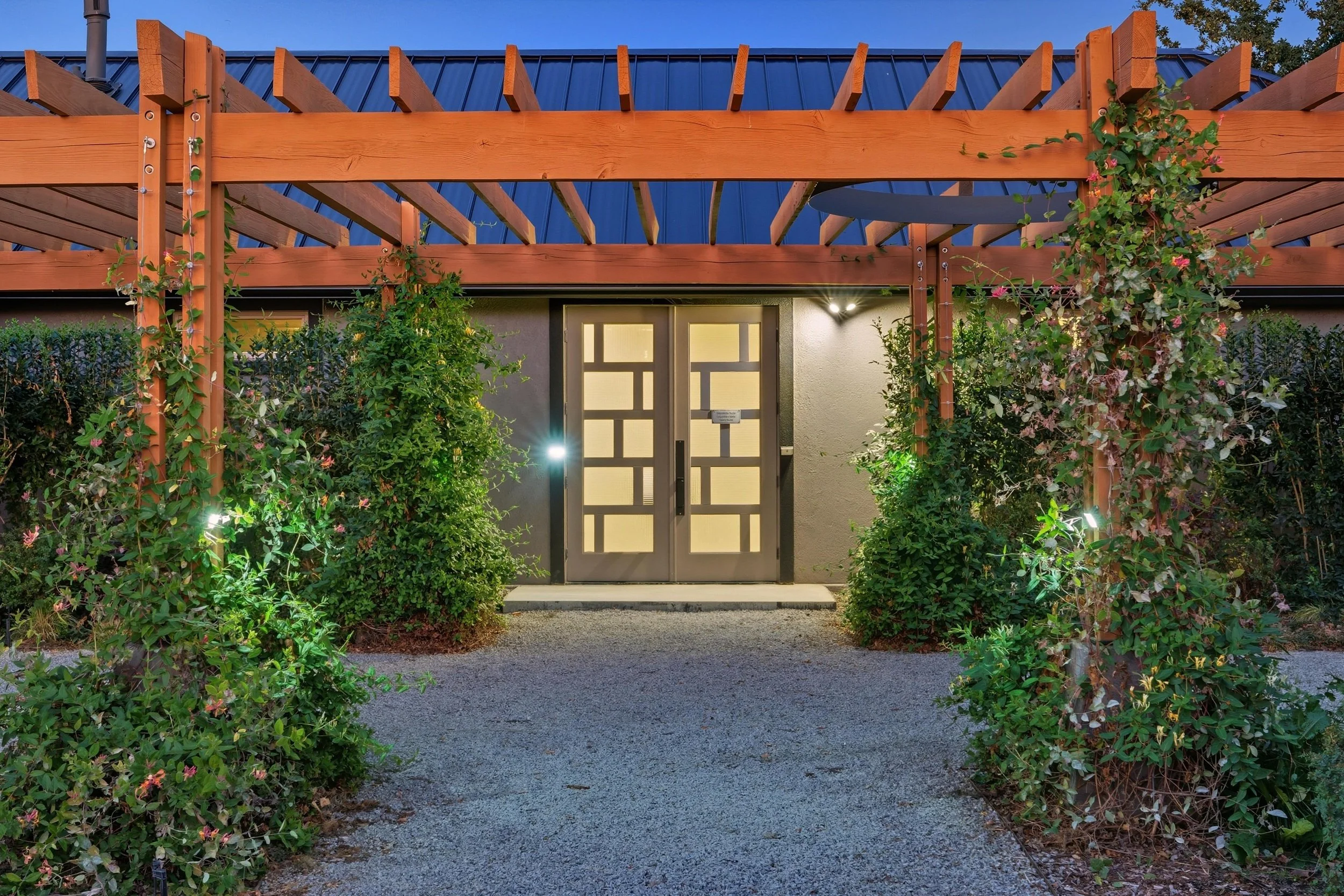Modern house entrance with double glass doors, illuminated by outdoor lights, surrounded by lush green bushes and a wooden pergola structure.
