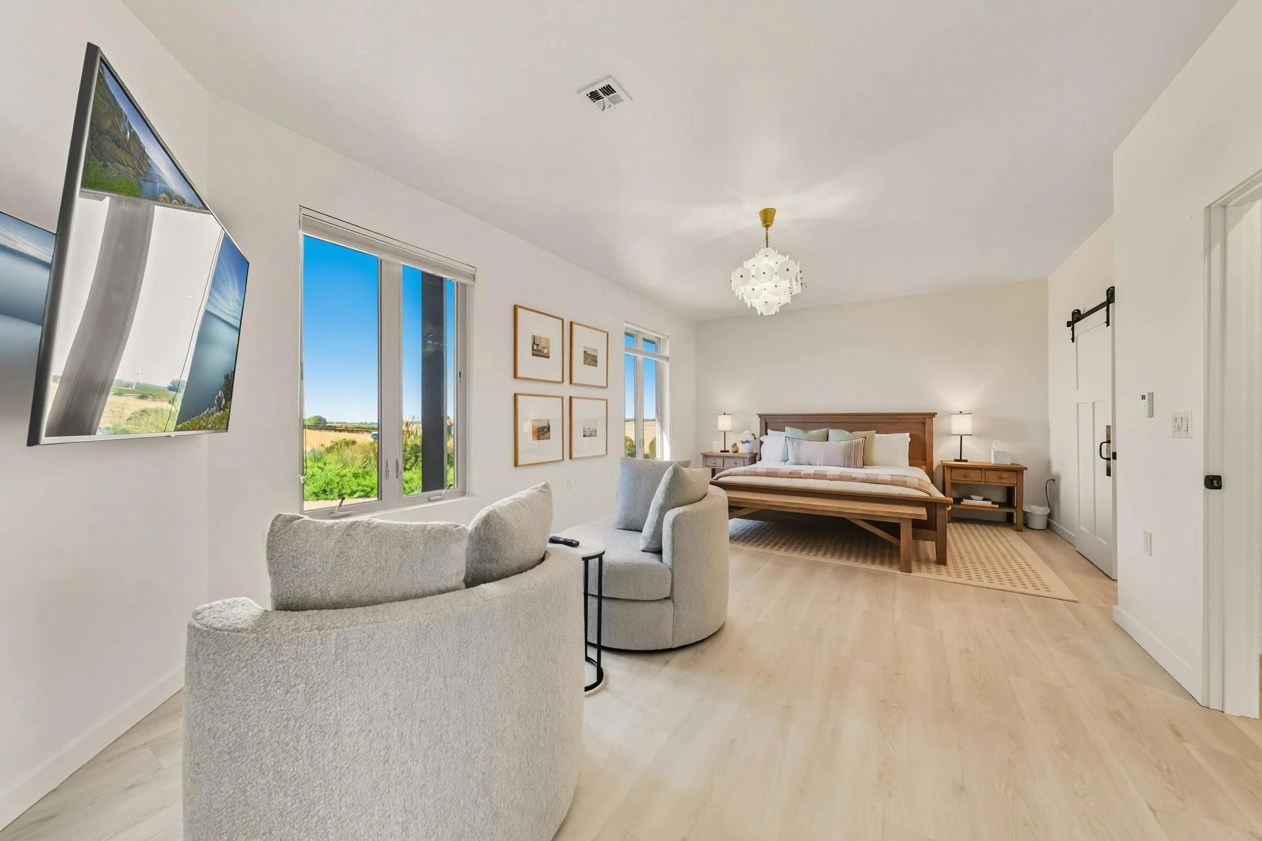 Bright, modern bedroom with white walls and light wood flooring, featuring a bed with wooden headboard, two nightstands with lamps, a small sitting area with a beige sofa and cushions, and a wall-mounted flat-screen TV. Large windows display a scenic