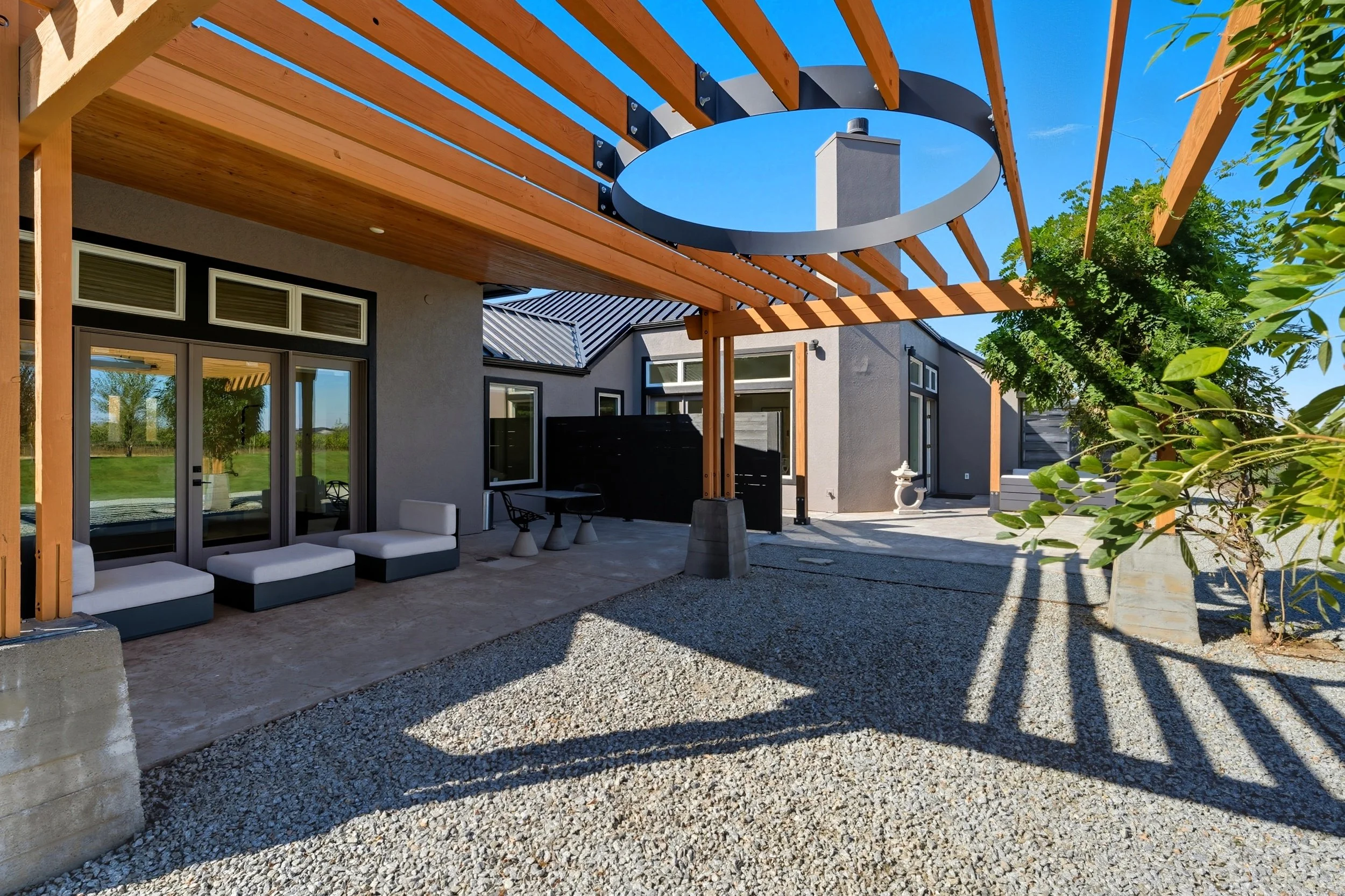 Modern backyard patio with gravel ground, outdoor seating, and a wooden pergola structure casting shadows. The house has large windows and a sliding glass door.