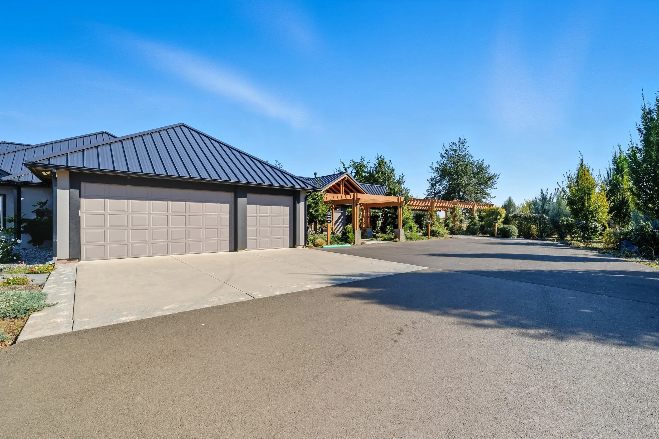 Modern residential house with a blue metal roof, gray garage doors, surrounded by trees and landscaping.