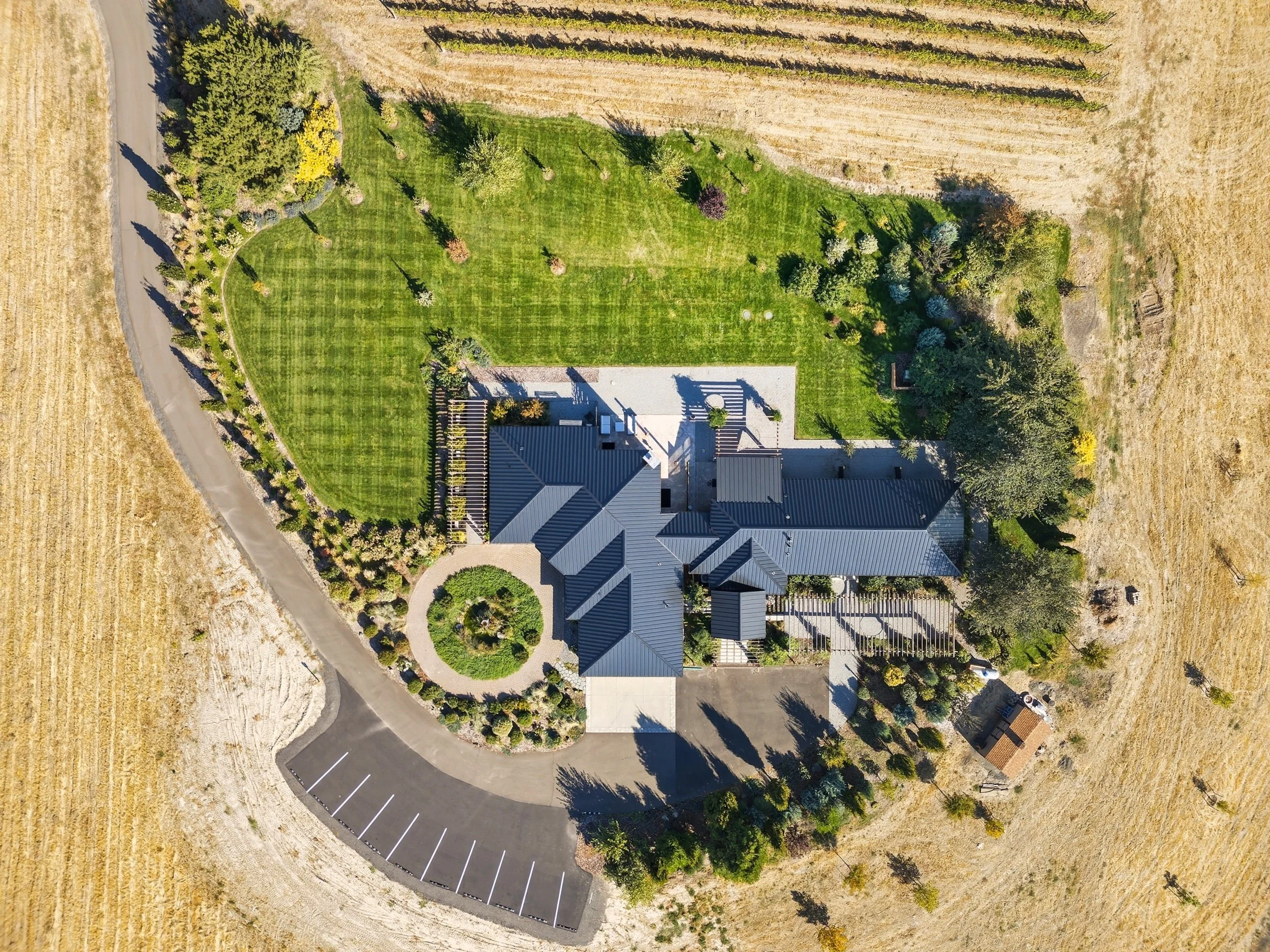 Aerial view of a house with a dark metal roof, surrounded by a green lawn, trees, and landscaped gardens, with a parking area and adjacent farmland.