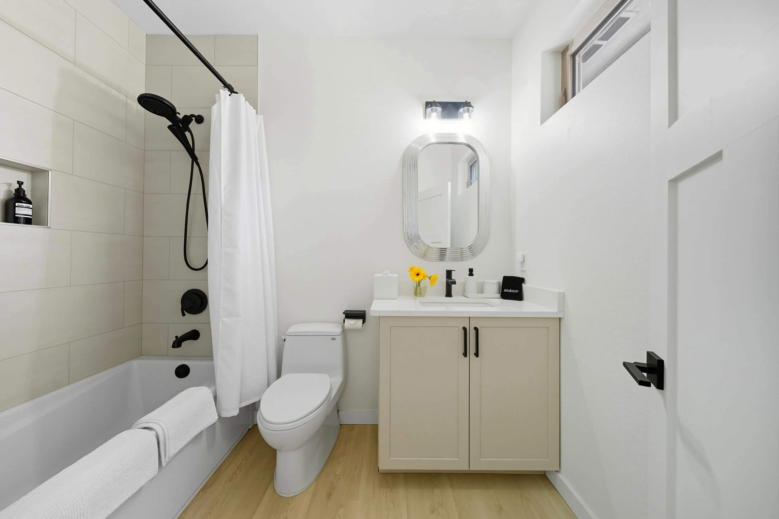 Modern bathroom with beige vanity, oval mirror, and a white bathtub with a white shower curtain. Small window and yellow flowers on the sink.