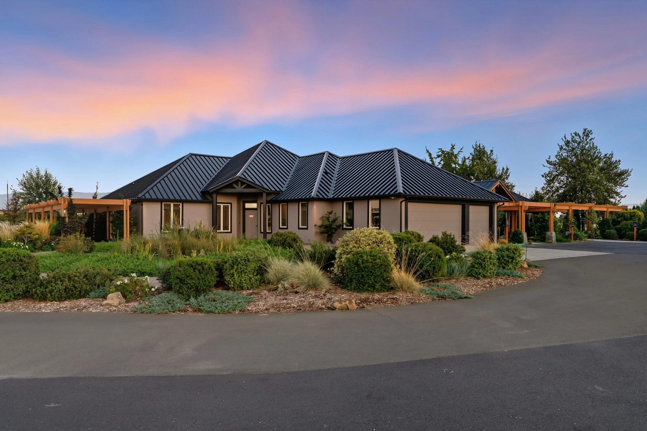 A modern house with a black metal roof and beige walls, surrounded by well-maintained landscaping and trees, under a colorful sunset sky.