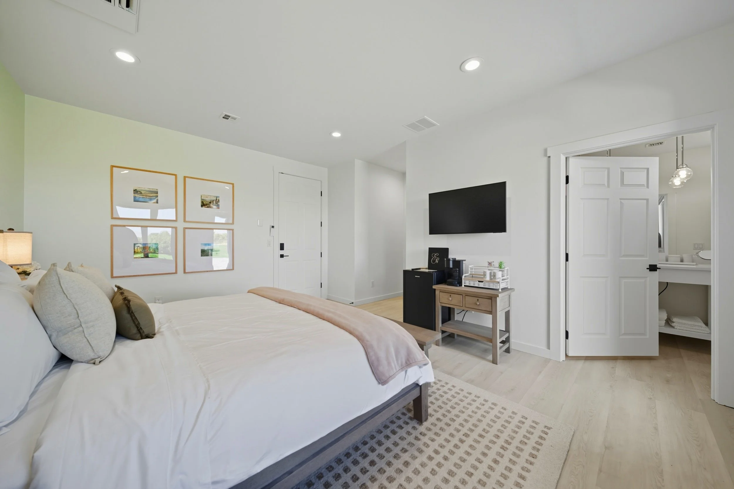 A modern bedroom with a large bed, four framed pictures on the wall, a small TV mounted on the wall, a dresser with a coffee machine, and a doorway leading to a bathroom.