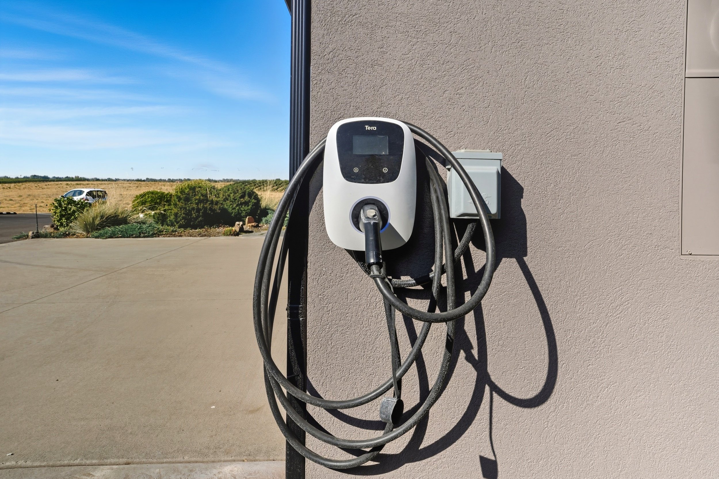 Electric vehicle charging station with an unplugged charging cable mounted on a beige exterior wall outside a building, with a parking lot, natural landscape, and blue sky in the background.