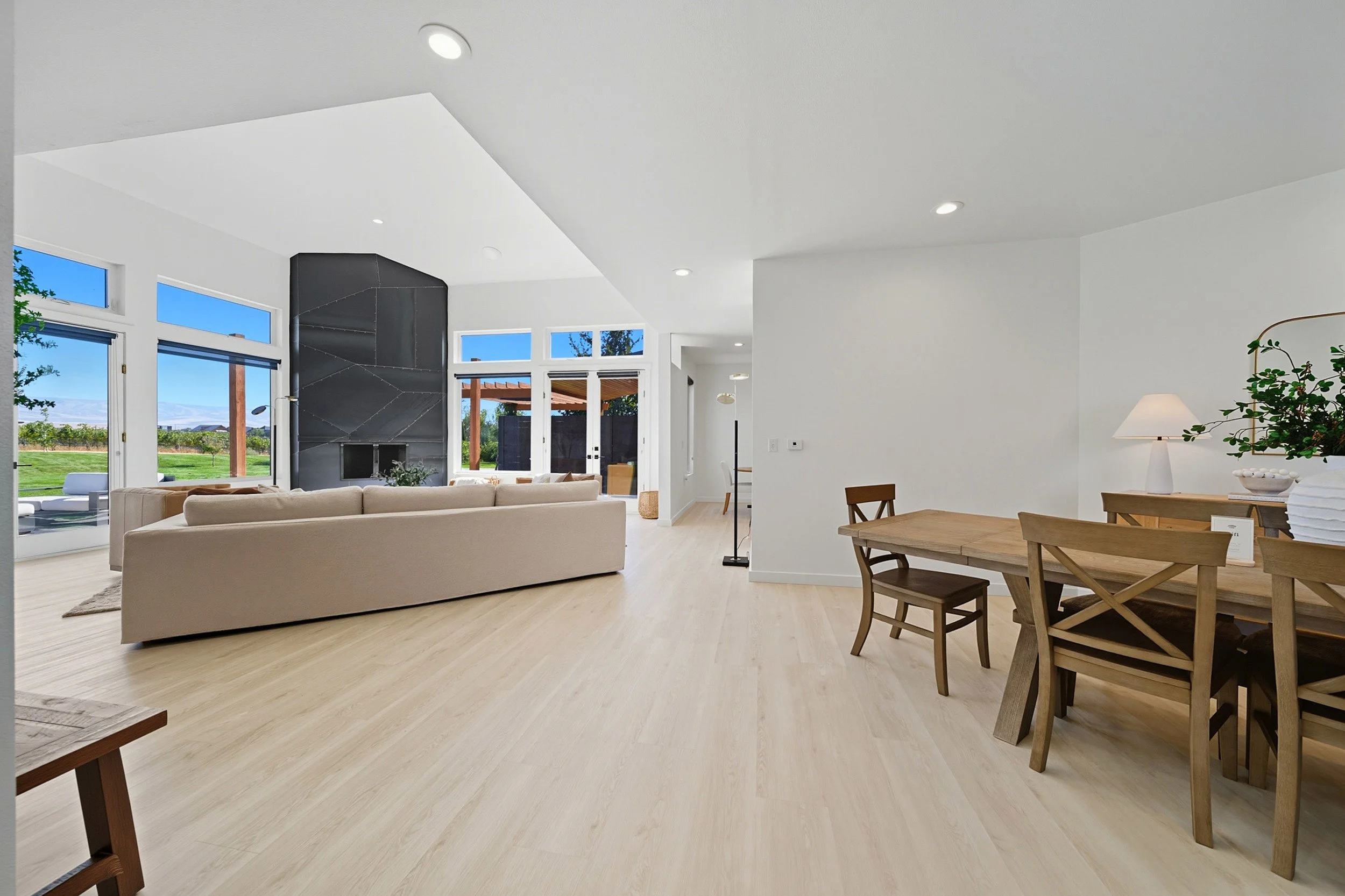 Spacious living room with large windows, white walls, light wood flooring, beige sofa, black fireplace, and wooden dining table with chairs, decorated with a lamp and potted plant.