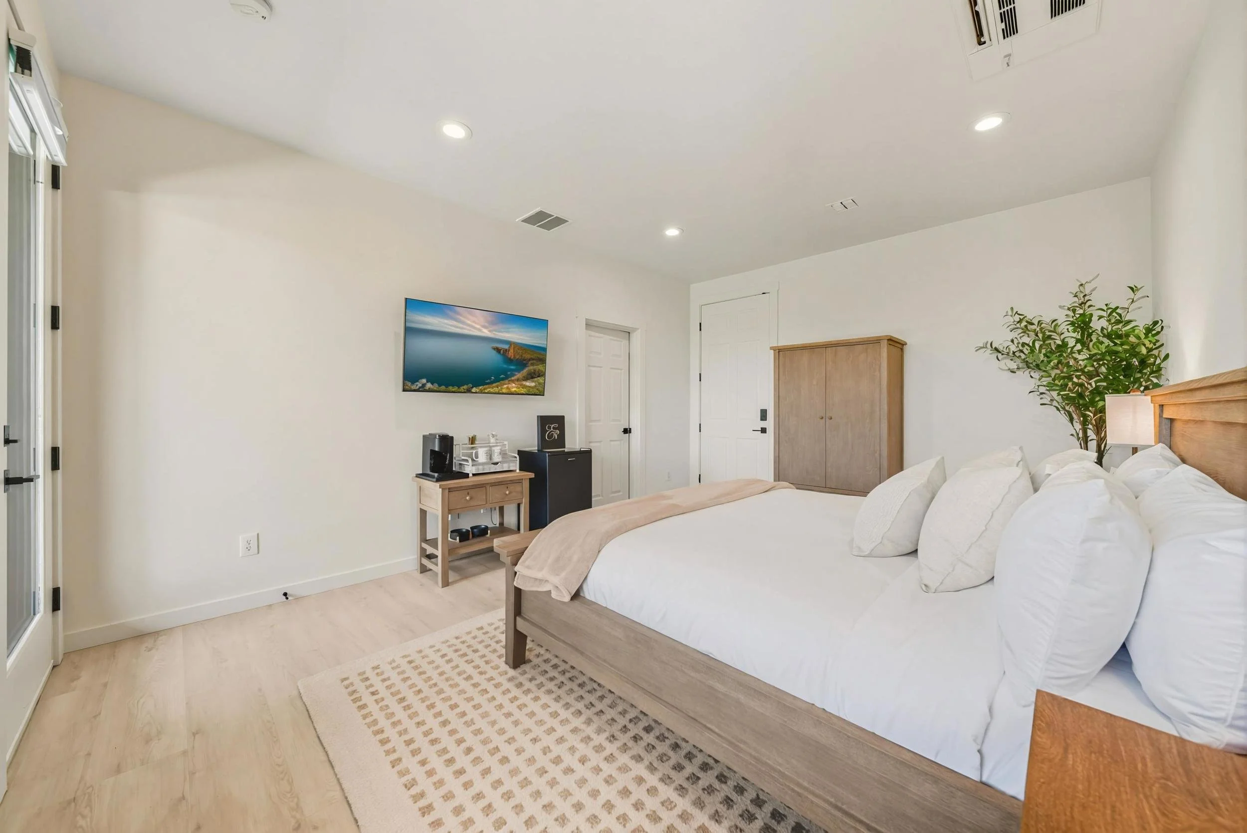 Modern bedroom with a large bed, white bedding, and several pillows. There is a wooden wardrobe, a small table with a coffee maker and snacks, a mounted television showing a coastal scene, a potted plant, and wall-mounted air conditioning vents. The 