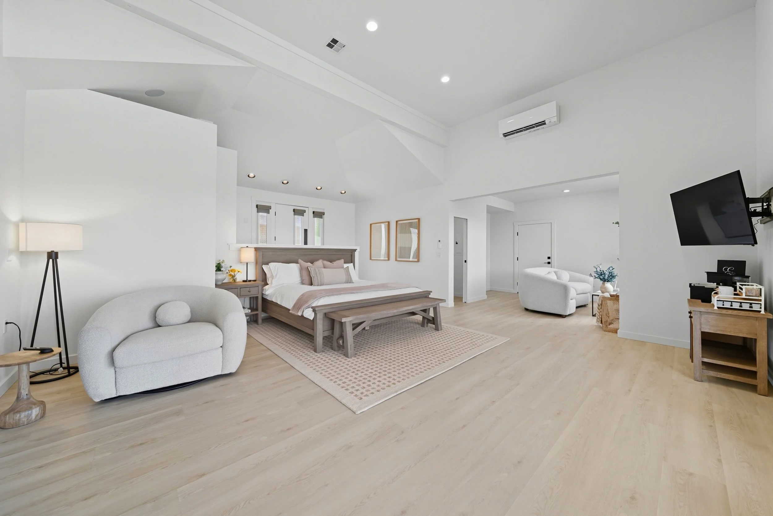 Spacious, bright bedroom with white walls, light wooden floors, a large bed with pillows, a bench, a small sofa, a wall-mounted TV, and minimal decor.