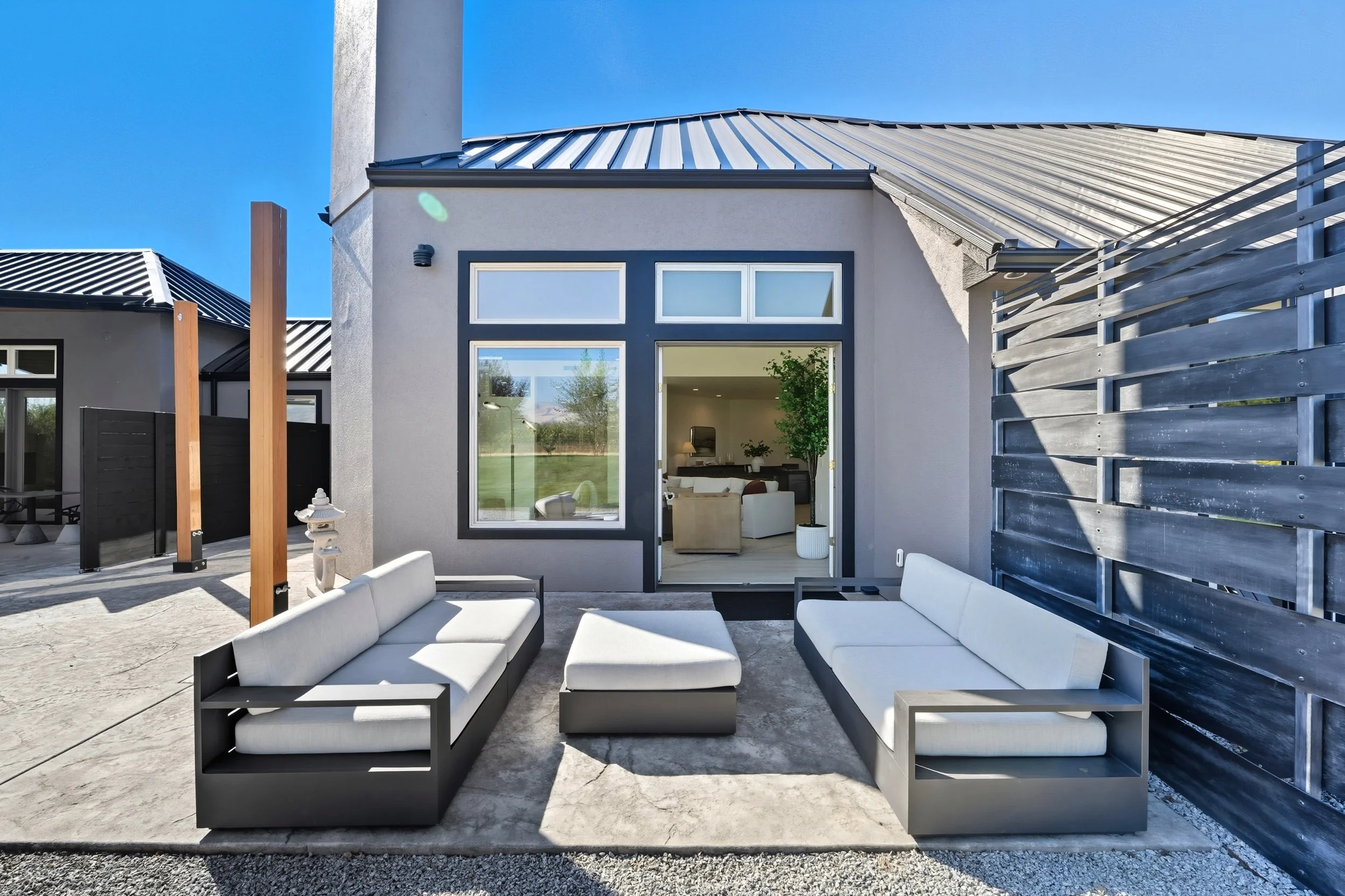 Modern outdoor patio with two white cushioned sofas and matching ottoman in front of a house with large windows and sliding door, enclosed by a wooden privacy screen, under a clear blue sky.