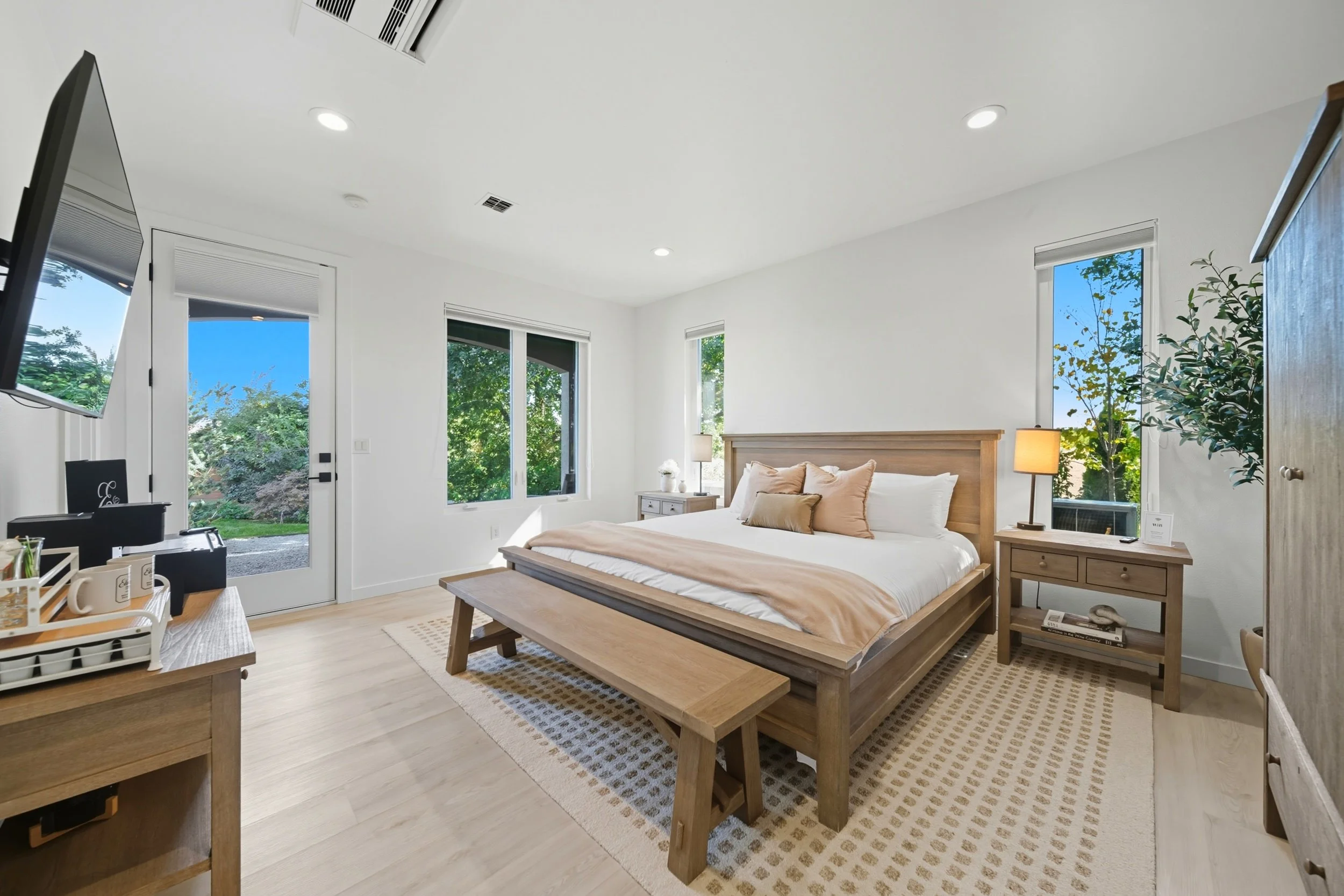 Bright bedroom with a wooden bed, beige bedding, and pillows, flanked by two small nightstands with lamps, large windows showing greenery outside, and a door leading outside.