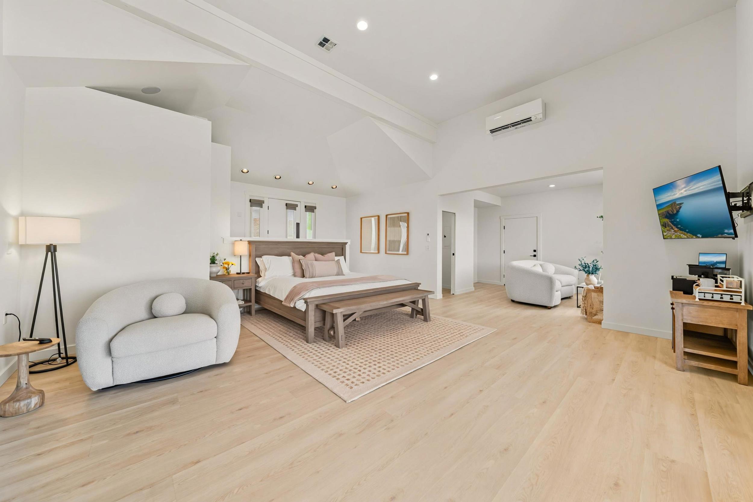 Spacious bedroom with white walls, light wood flooring, a bed with neutral-colored bedding, a small bench at the foot of the bed, and a sitting area with a white armchair and a TV mounted on the wall.