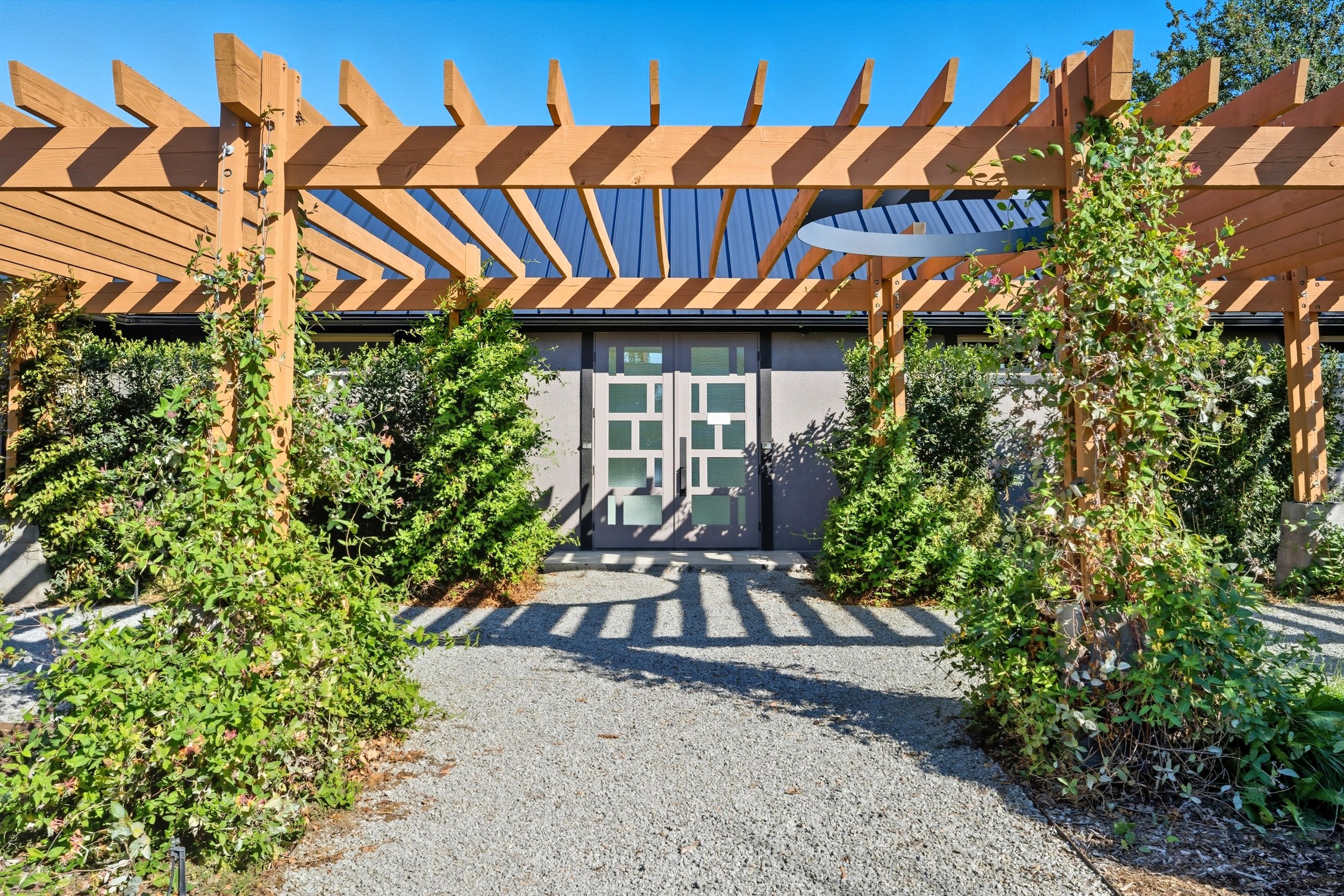 A modern outdoor garden space with a wooden pergola, green bushes, a concrete wall with a patterned green door, and shadows cast by the pergola in bright sunlight.