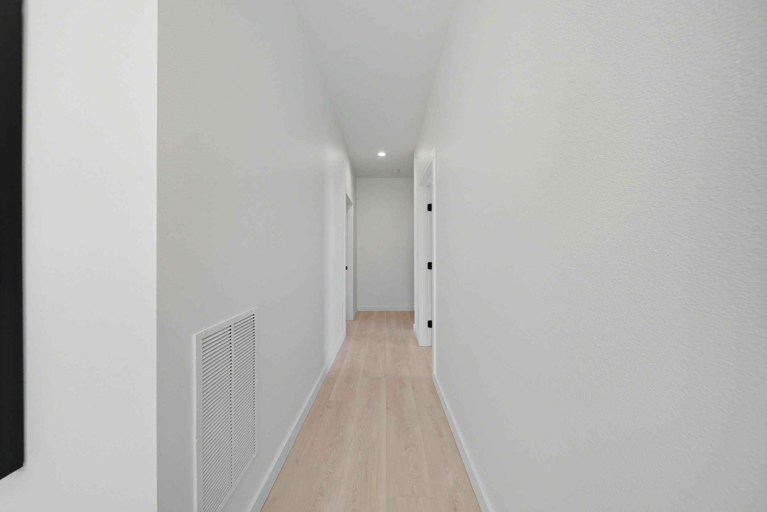 Empty white hallway with light wood flooring, a vent on the lower part of the left wall, and three closed white doors on the right side.